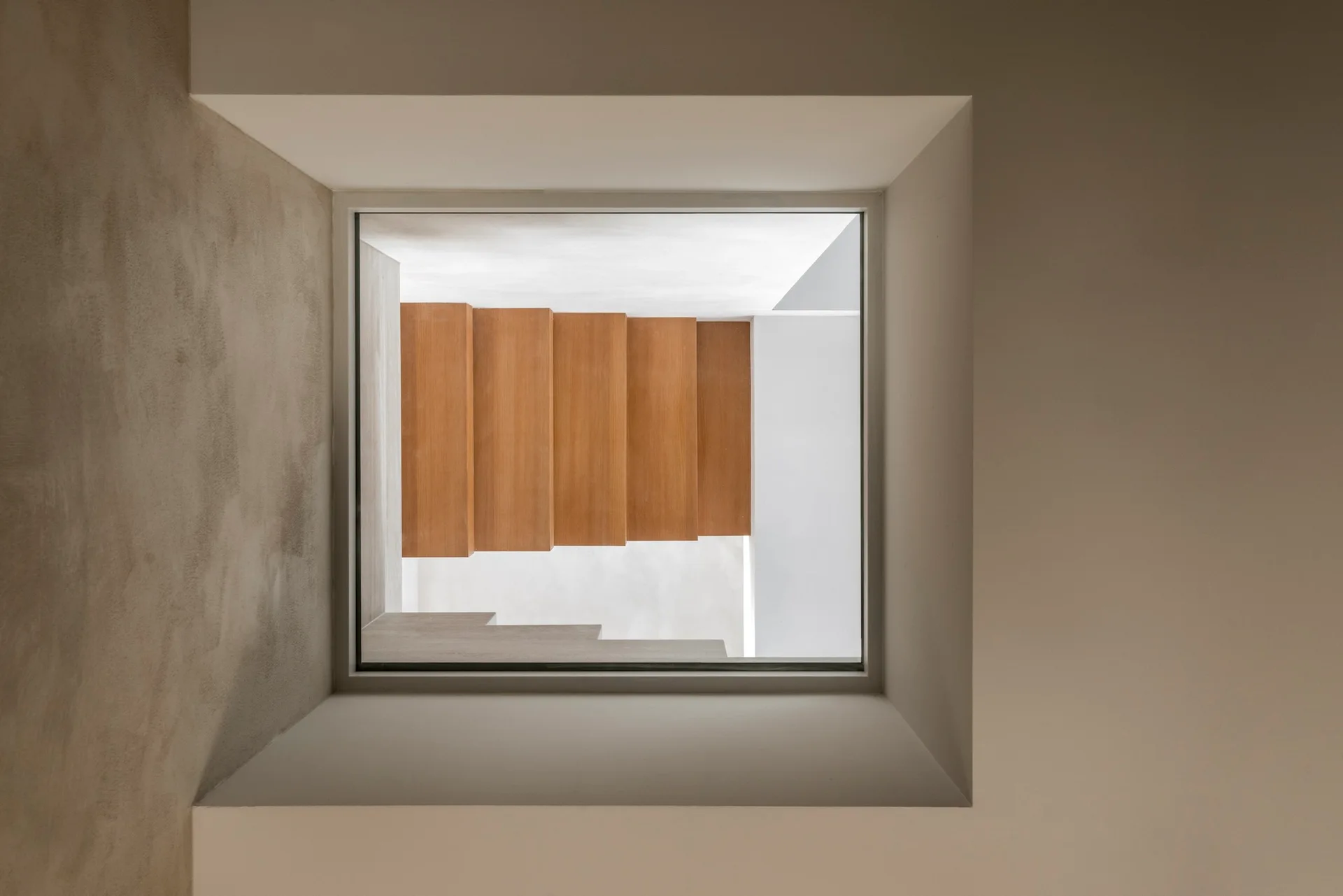 A top-down view through a square recessed ceiling opening looks down onto minimalist wooden stair treads surrounded by smooth white walls.