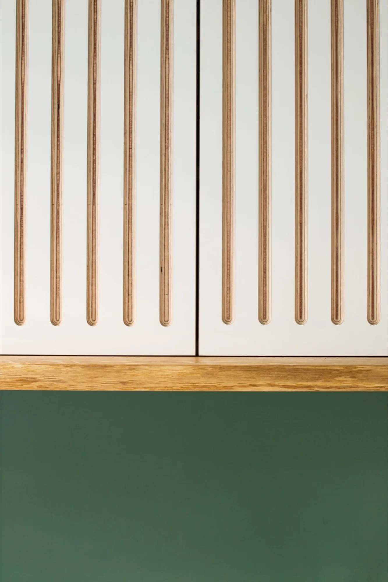 White cabinet doors featuring vertical wood slat details sit above a natural wood shelf and a solid green painted wall.