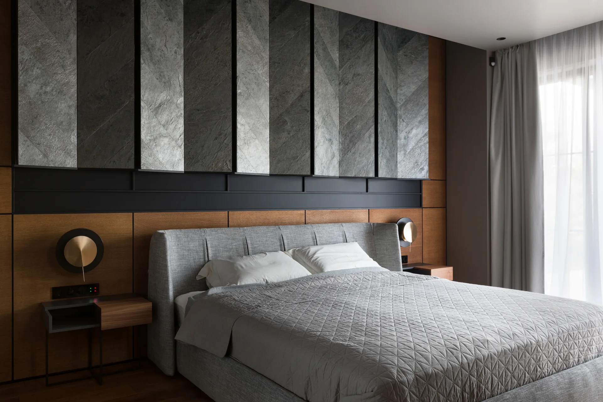 A modern bedroom features a gray upholstered bed set against a feature wall of warm wood panels and vertical gray textured stone slabs.