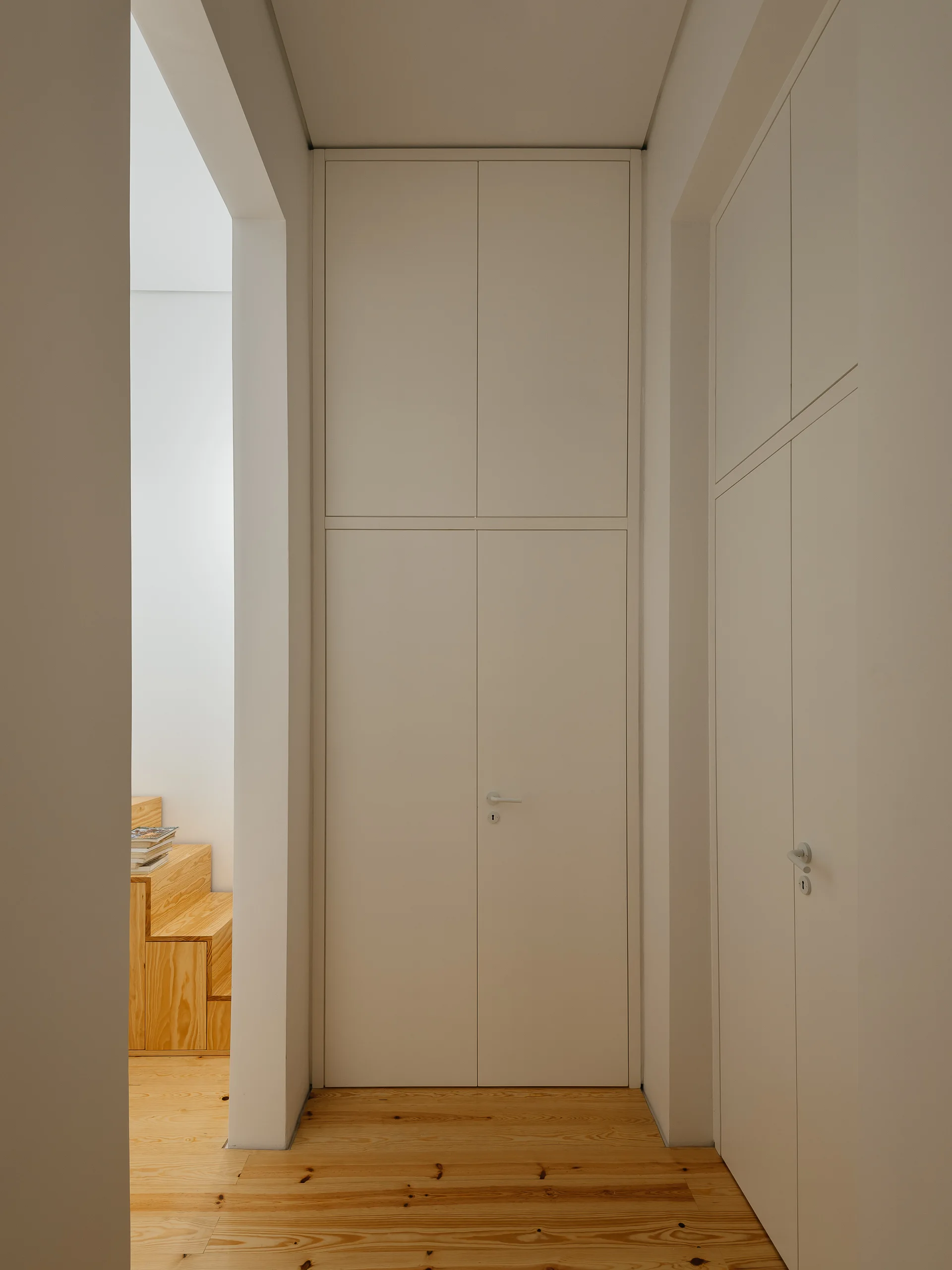 A minimalist hallway features flush floor-to-ceiling white cabinets, light wood flooring, and an open doorway leading to a wooden staircase.