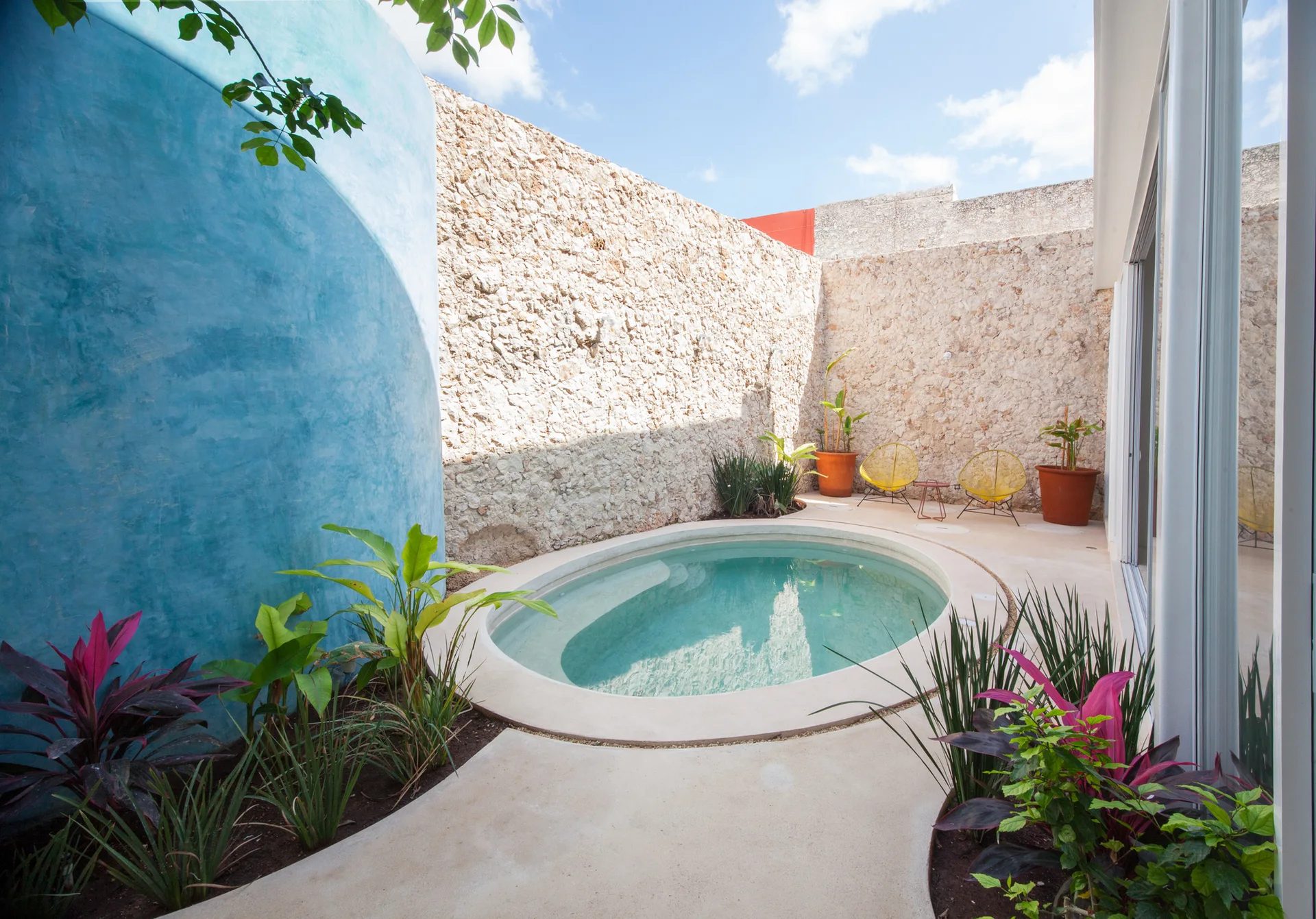 An oval swimming pool in a concrete courtyard patio enclosed by a high stone wall and a curved blue plaster wall with tropical plants.