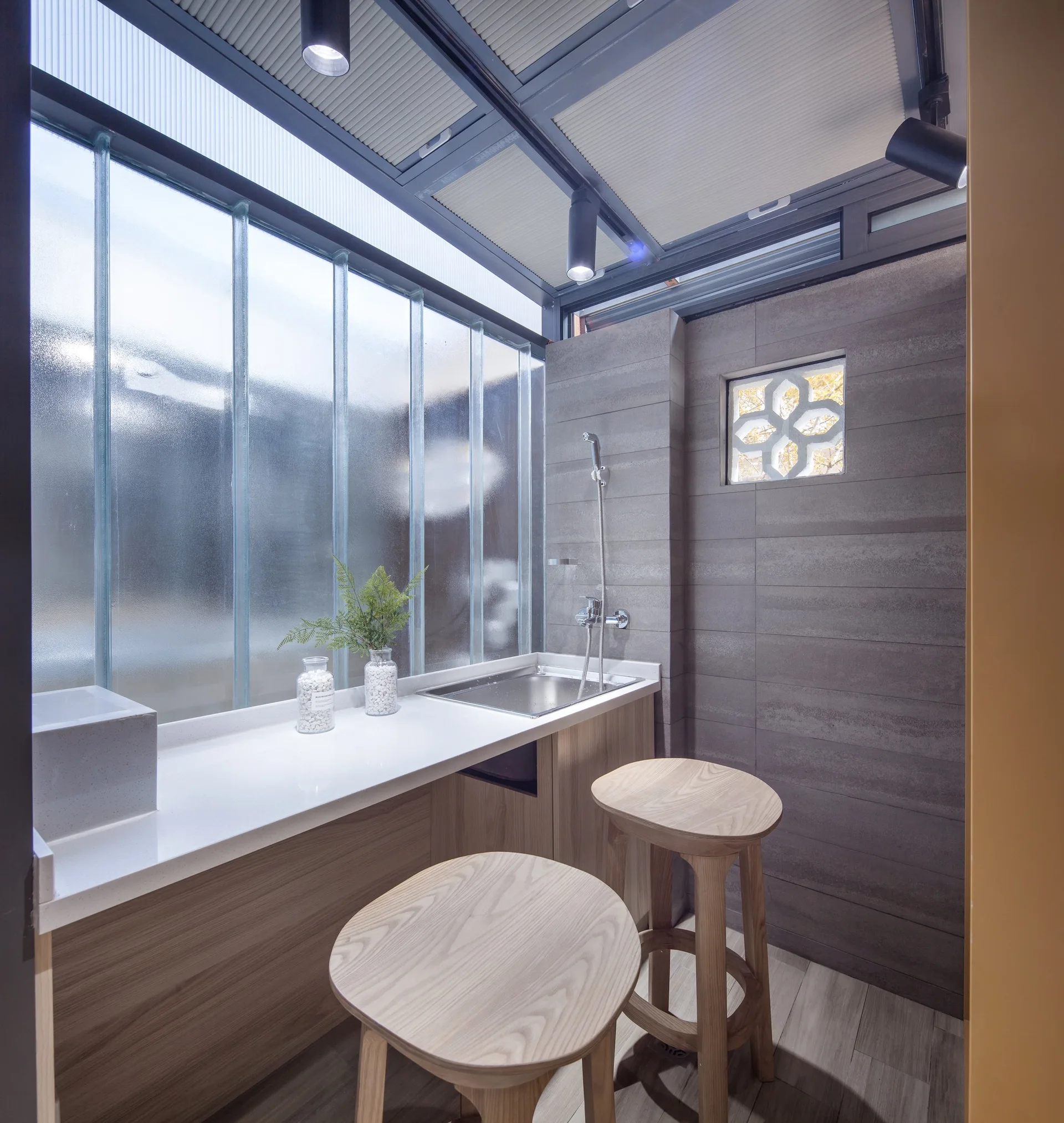 A compact utility room features wooden stools, a metal sink on a white countertop, a frosted glass wall, and grey concrete panels.