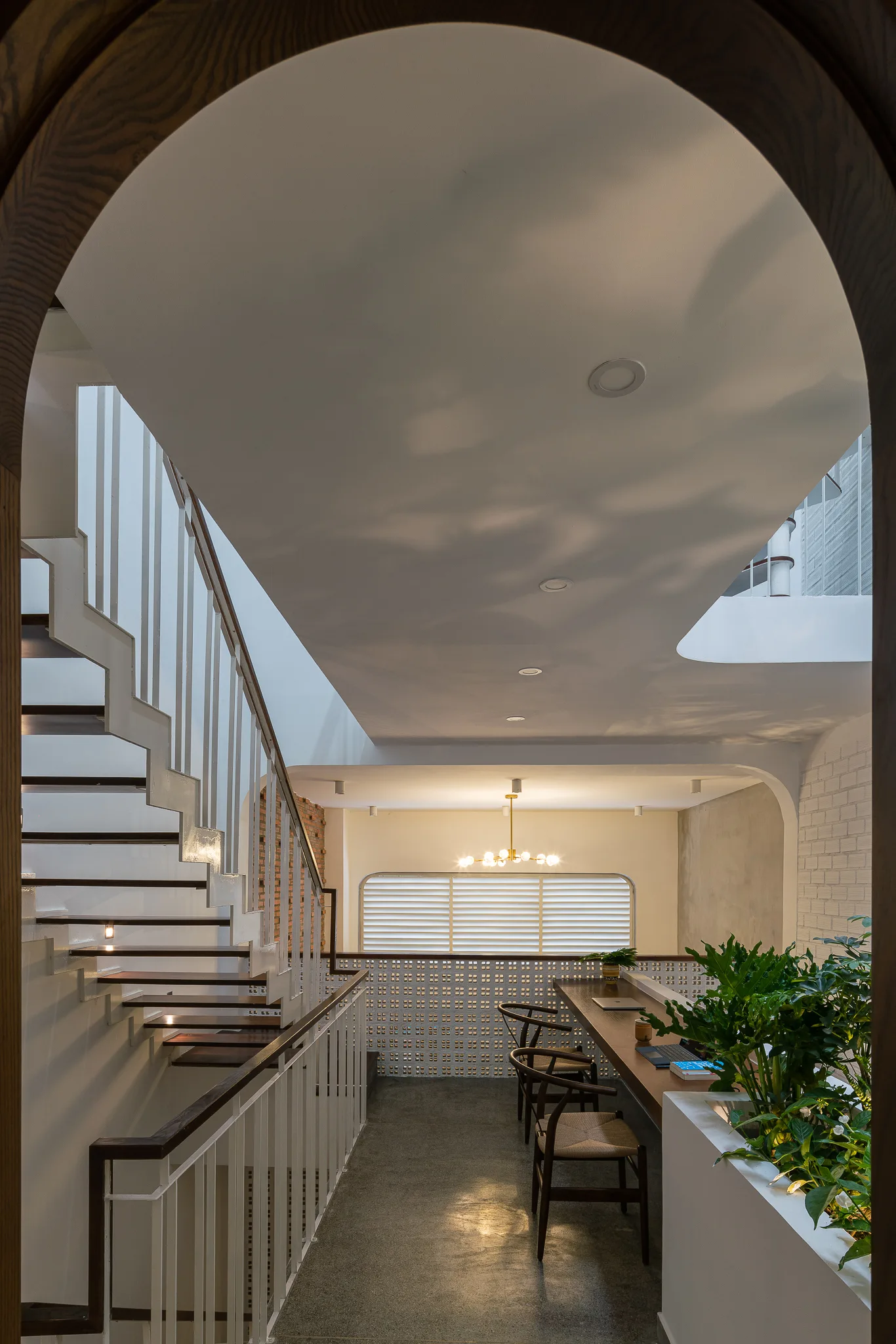 A modern home interior with a wooden staircase and a mezzanine workspace featuring a long desk and breeze-block screen wall, seen through a wooden archway.