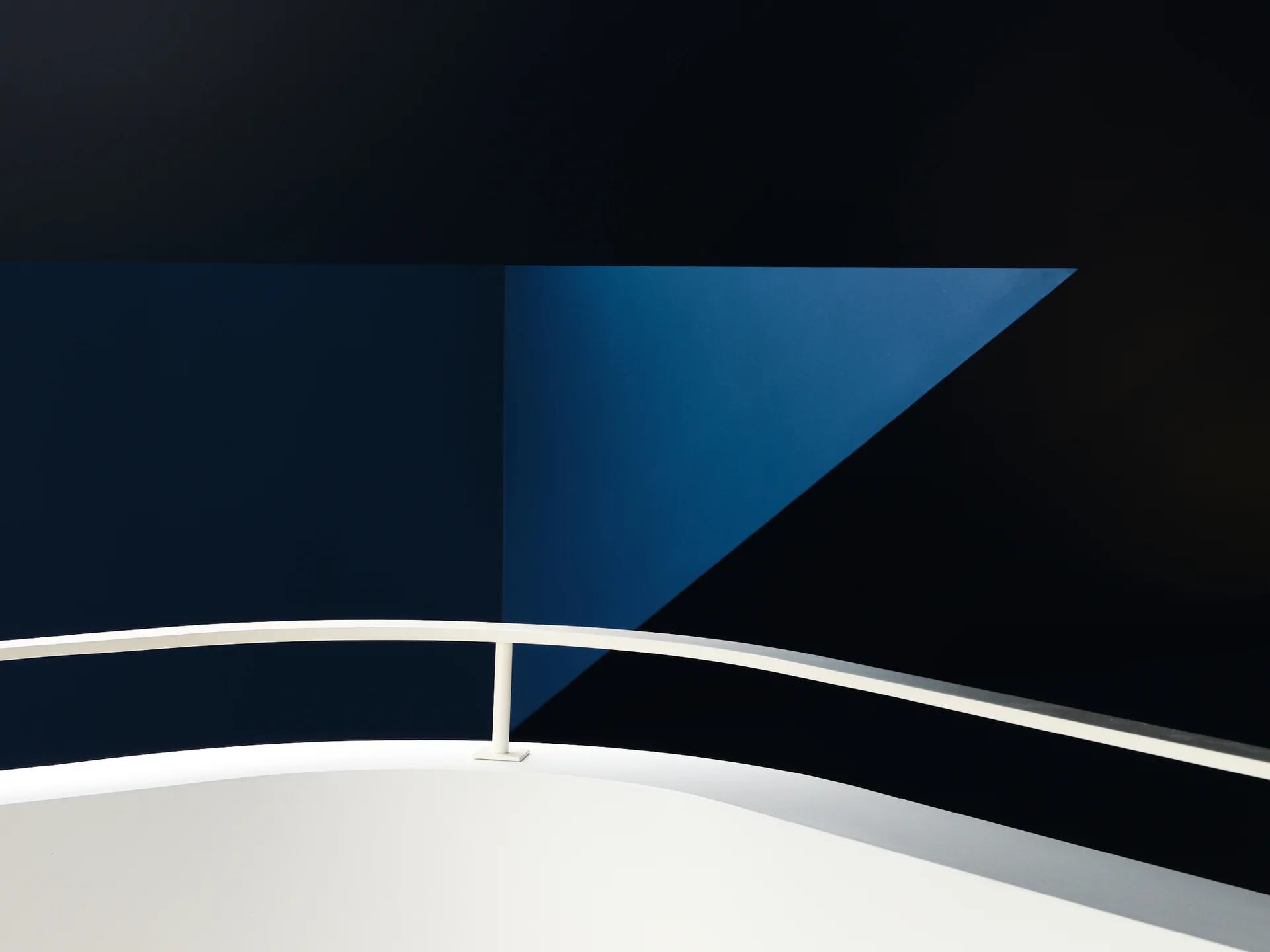 A curved white metal railing on a landing set against a high-contrast background of intersecting black and blue matte geometric painted walls.