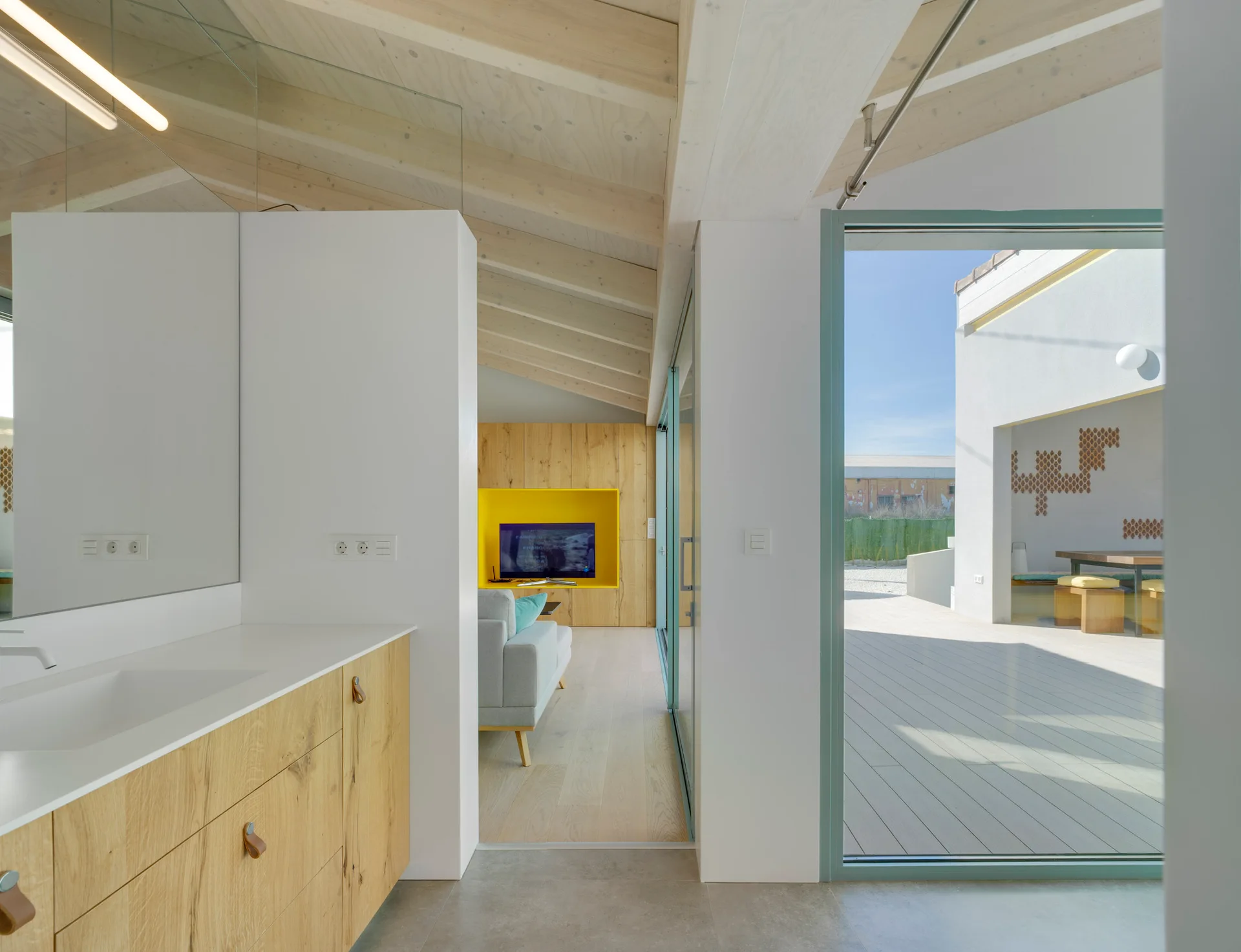 A modern interior showing a wooden bathroom vanity transitioning to a living area with a yellow TV niche and an outdoor wood-decked patio.