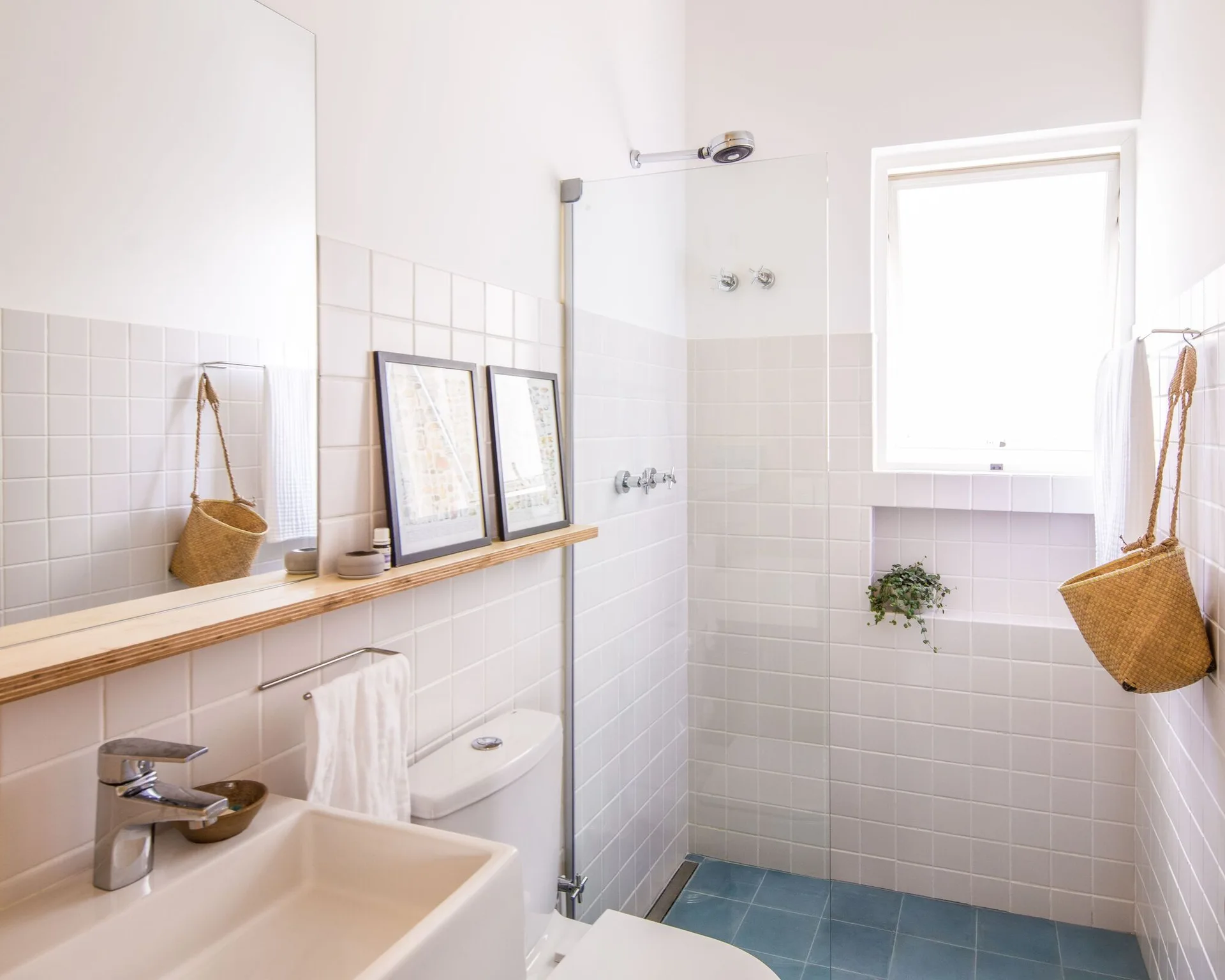 A bright bathroom features white grid-tiled walls, a blue tiled floor, a glass shower partition, and a slim wood shelf below a large mirror.