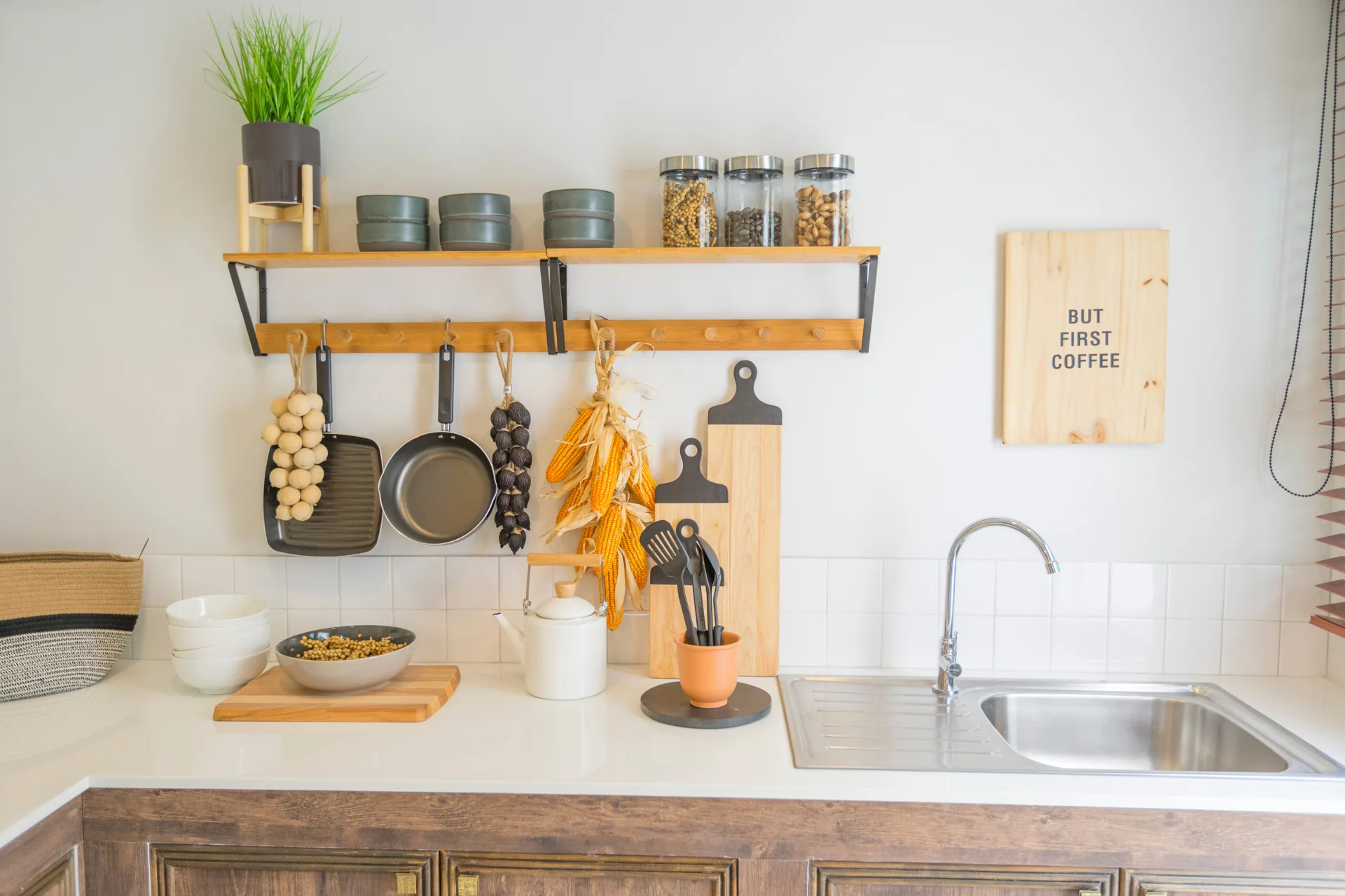 A white tiled kitchen counter features a stainless steel sink, a wooden shelf, a peg rail with hanging pans, and cutting boards.