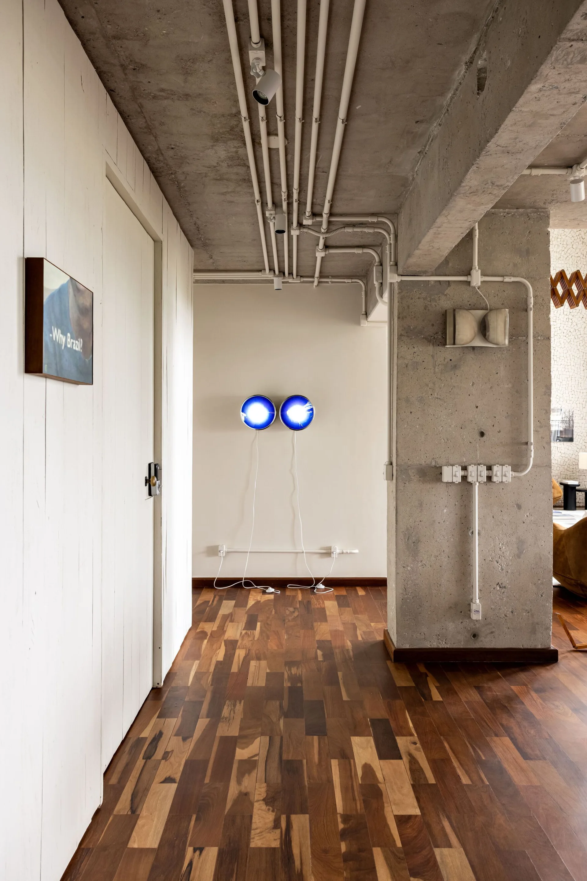 A residential hallway features warm wood plank flooring, a raw concrete column and ceiling, white paneled walls, and two glowing blue circular sconces.