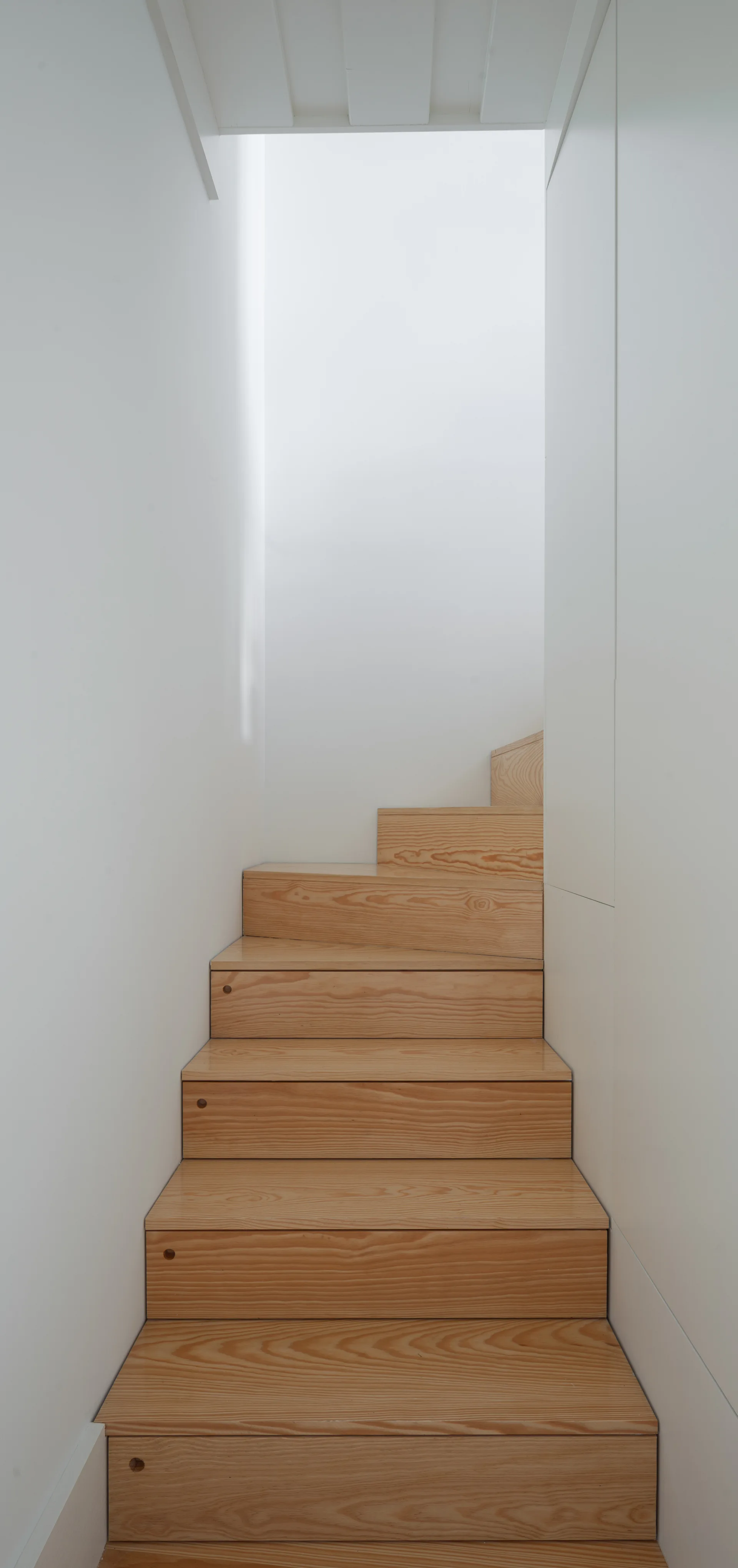 A narrow wooden staircase with prominent grain rises between smooth matte white walls under natural light.