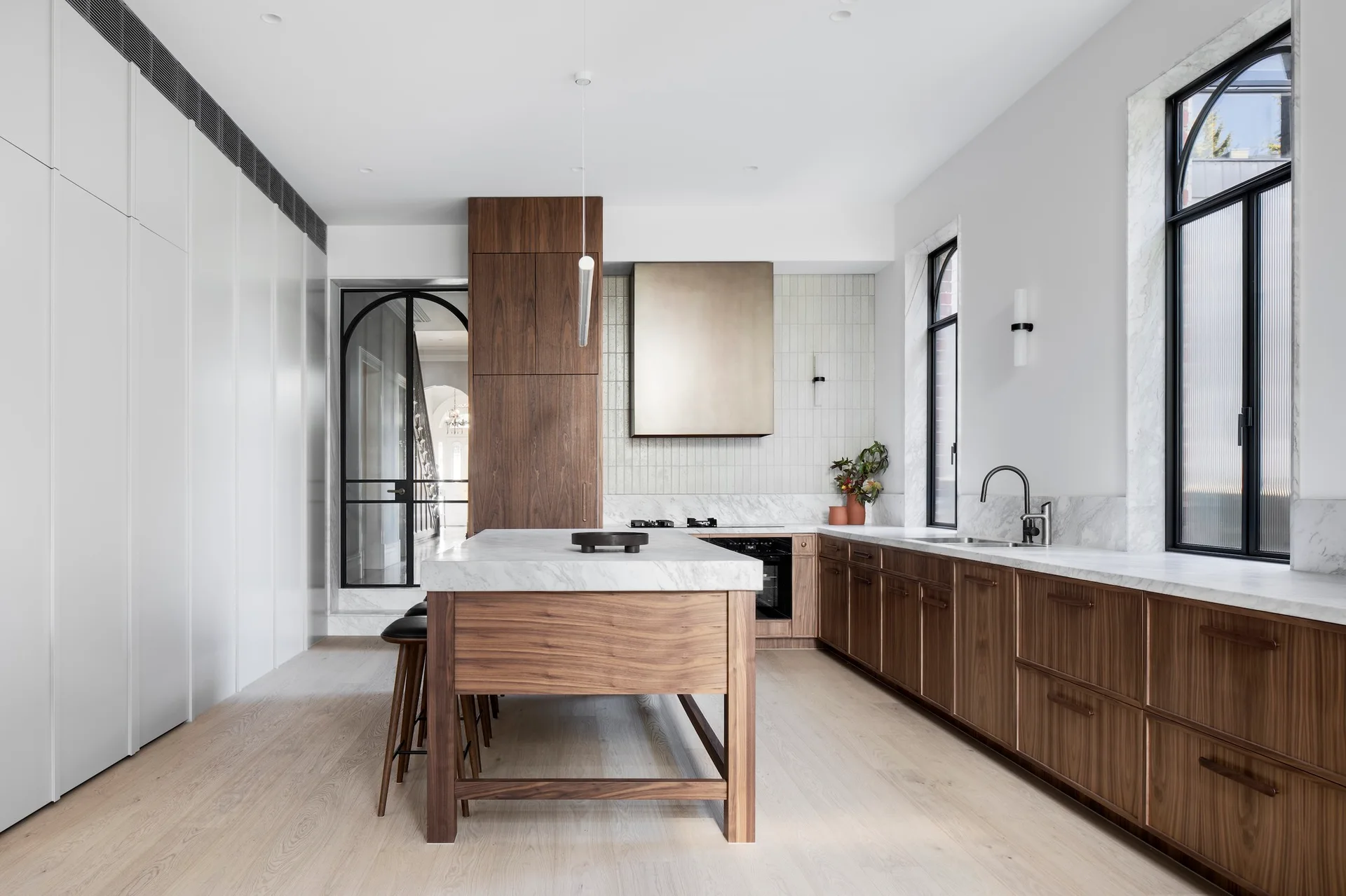 A contemporary kitchen features dark walnut cabinetry, a marble-topped island, arched black metal windows, and a metallic range hood.