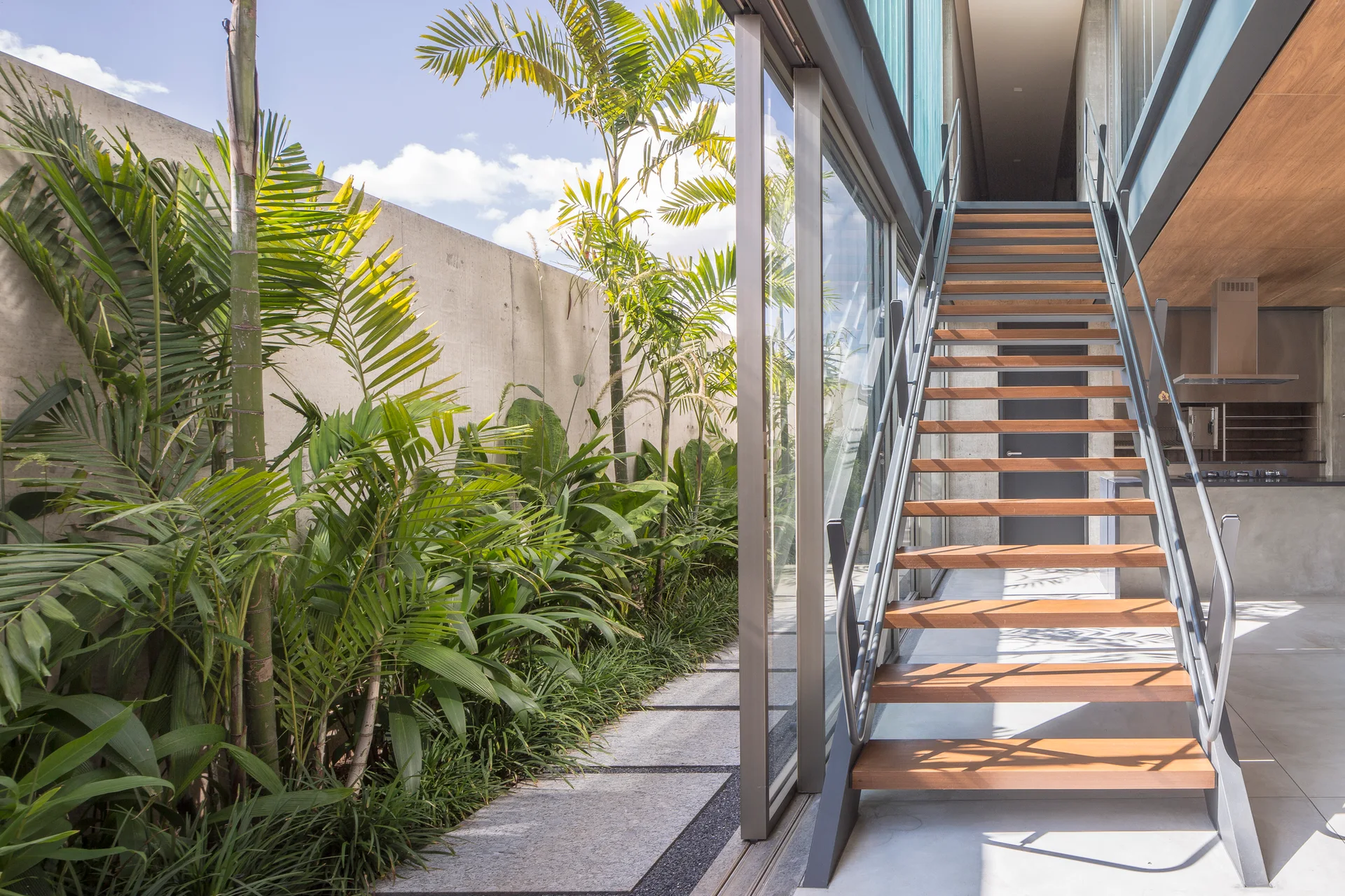 A modern open-riser wooden staircase sits next to a sliding glass door opening to a narrow tropical garden with palm trees and a concrete wall.
