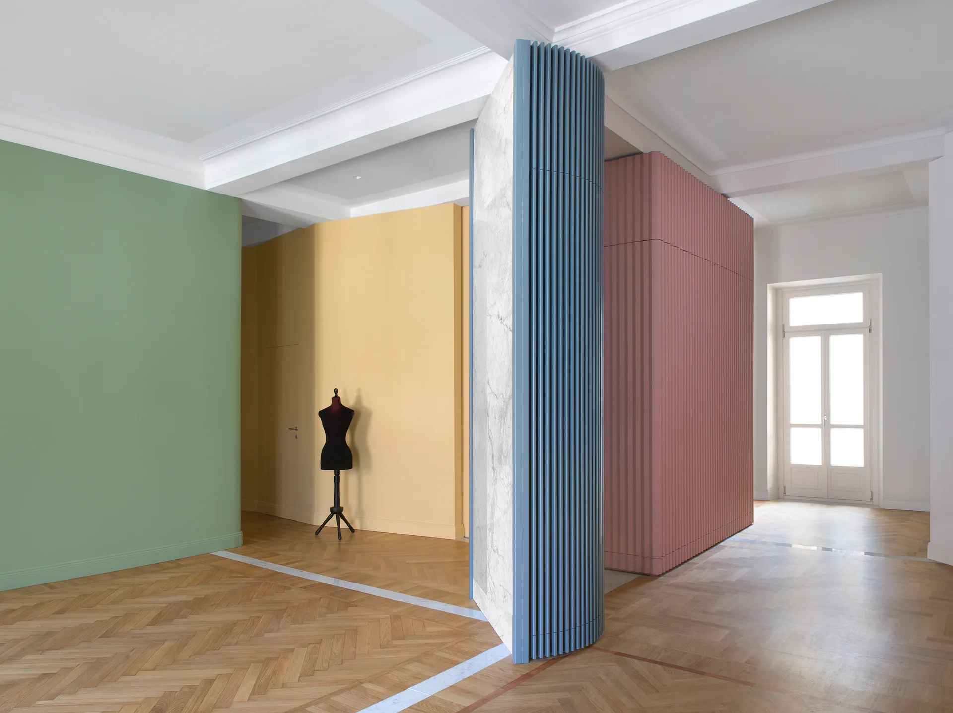 A spacious room features a central multi-colored sculptural partition with blue and pink fluted paneling over light brown herringbone hardwood floors.