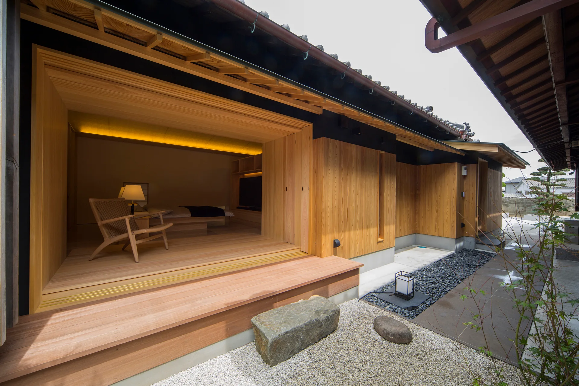 A modern Japanese-style bedroom with a woven armchair opens onto a wooden engawa porch next to a gravel patio with a stone bench.