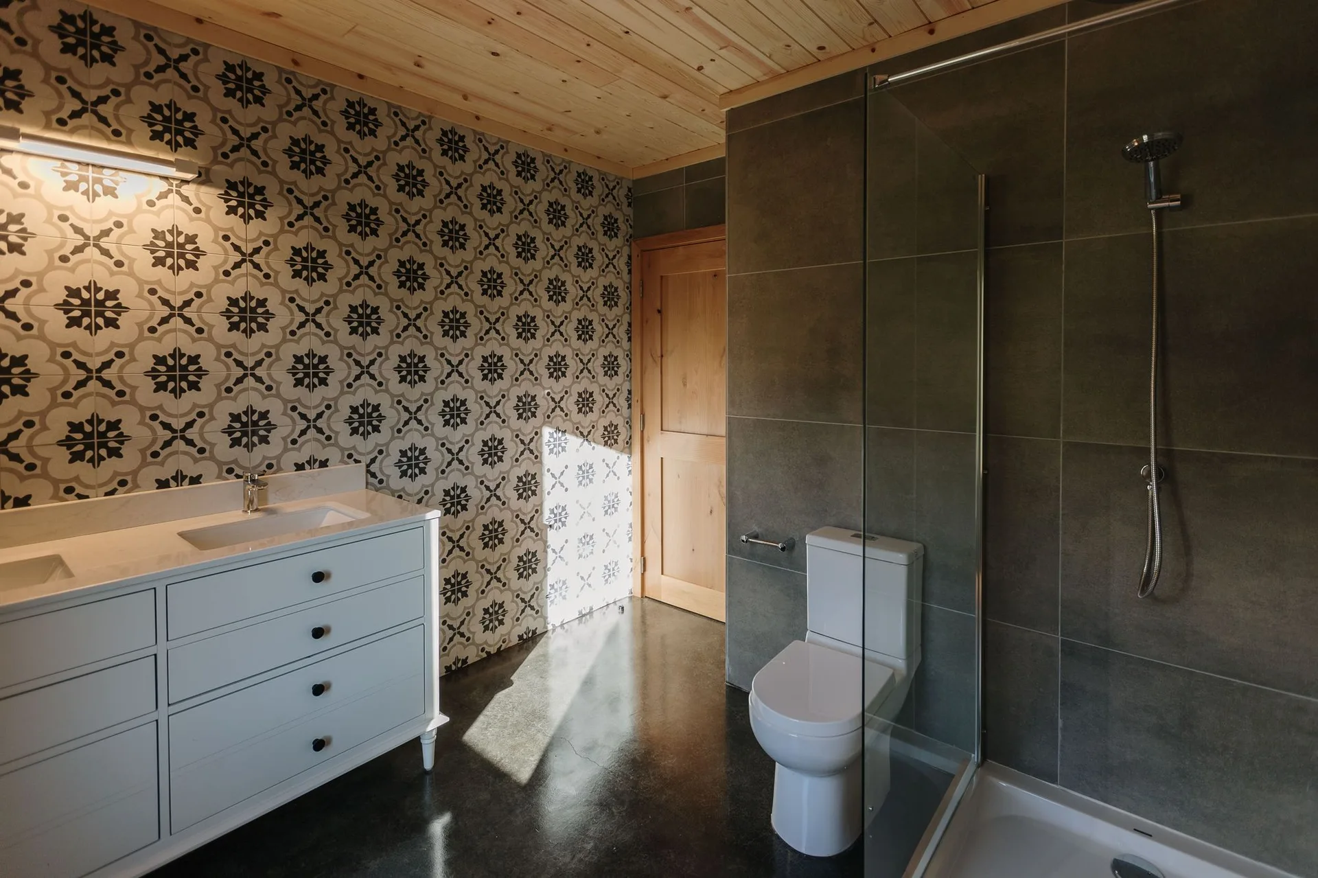 A modern bathroom features a white double vanity next to a geometric patterned wall, a light wood plank ceiling, and a grey tiled shower.