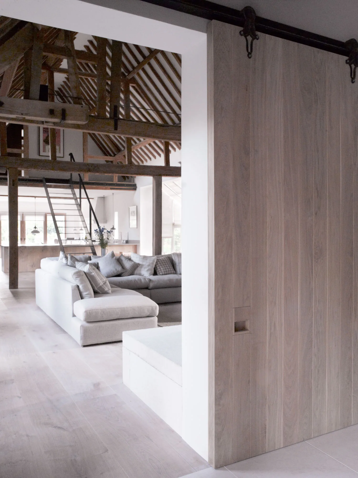 An open-plan living room featuring a grey sectional sofa, vaulted ceilings with exposed timber trusses, and a large wooden sliding barn door.