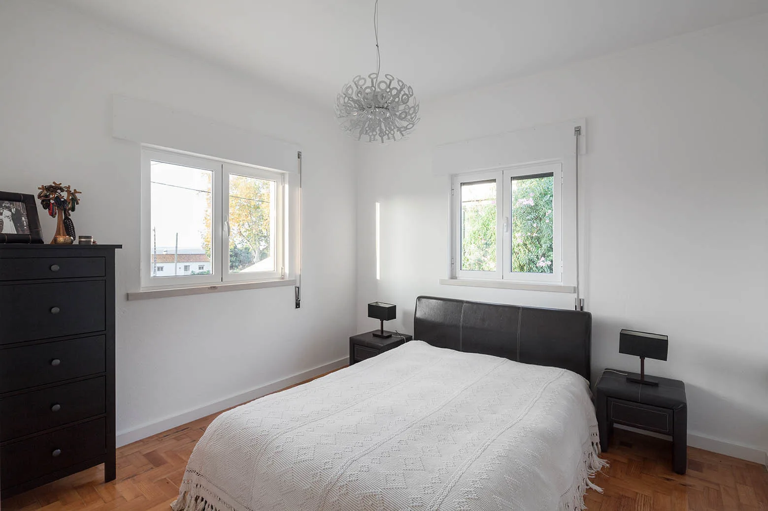 A modern bedroom features a black headboard bed, two matching black nightstands, a tall dresser, parquet flooring, and two white-framed windows.