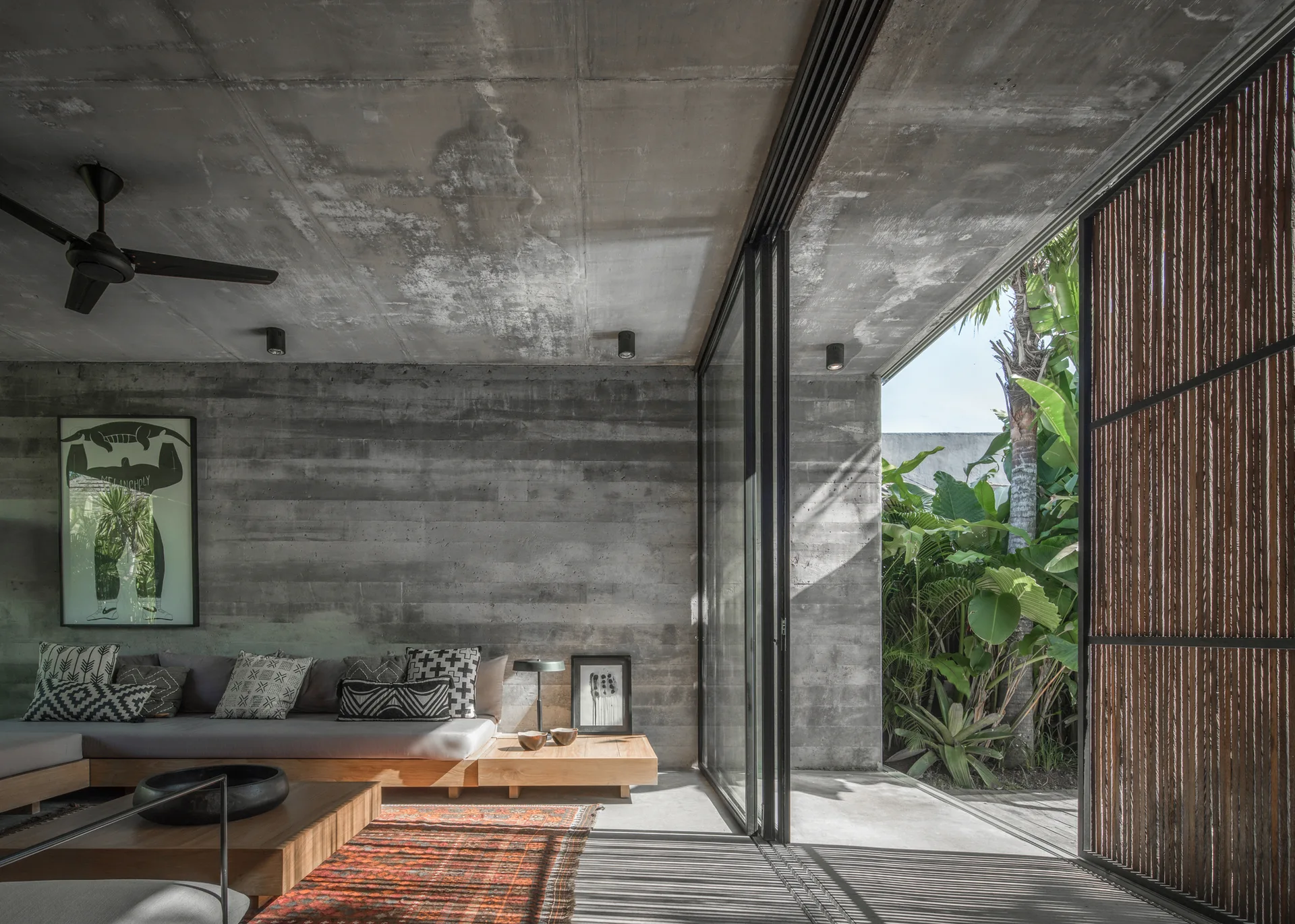 A living room with board-formed concrete walls, a low wooden sectional sofa, and a large sliding glass door opening to a lush tropical garden.