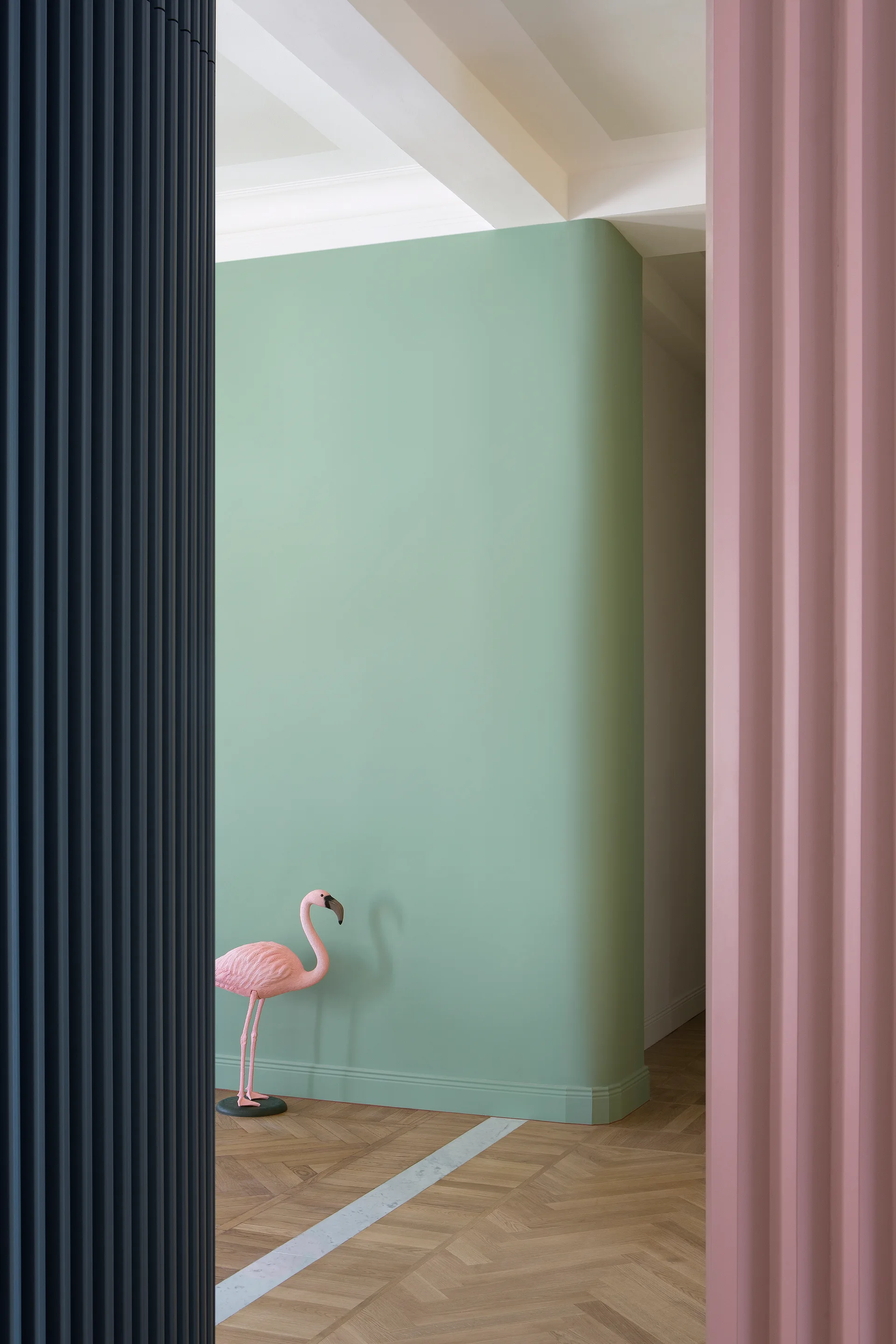 A view through dark blue and pink fluted columns reveals a curved mint green wall and a pink flamingo on herringbone wood floors.