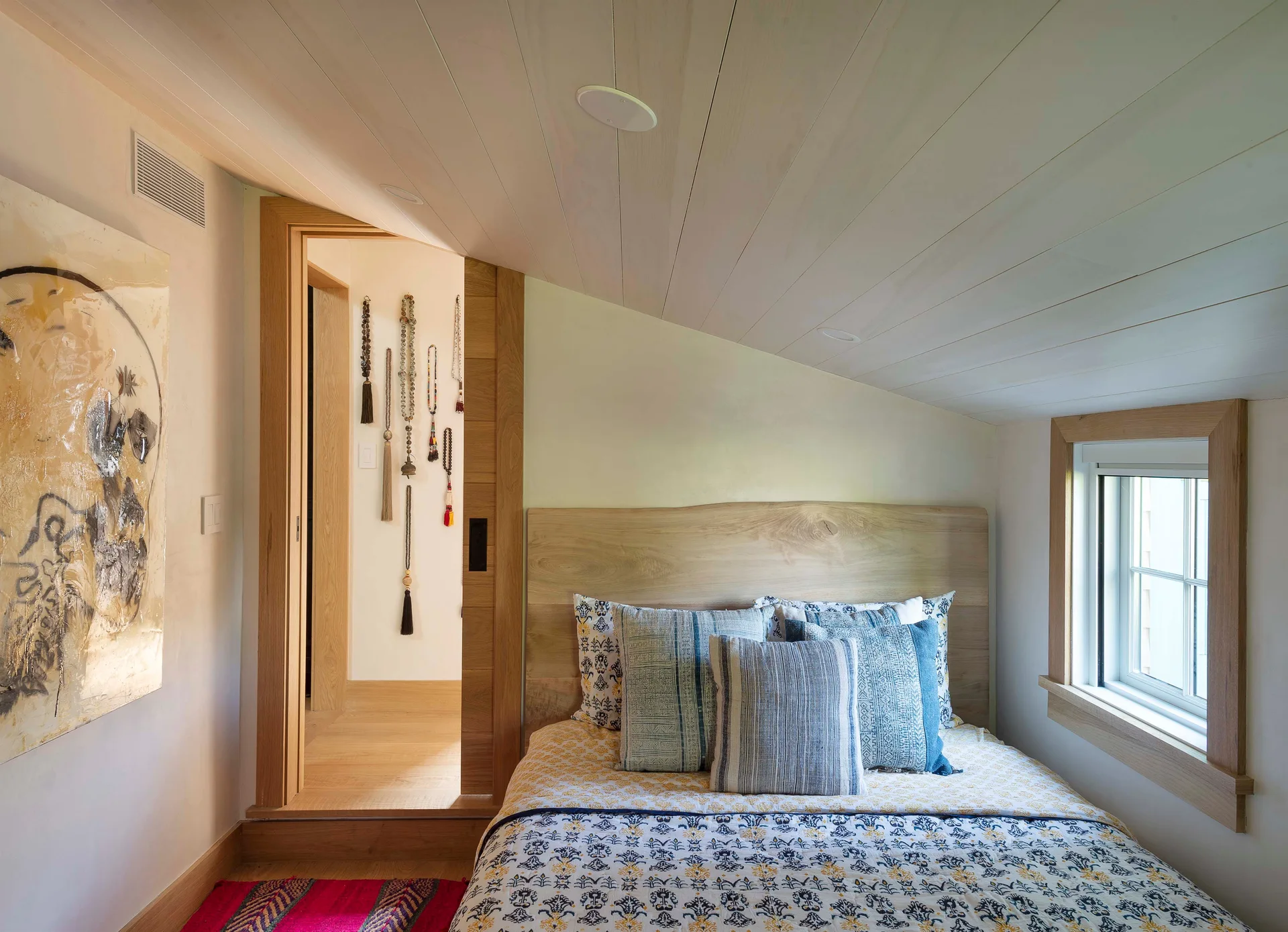 A compact attic bedroom features a bed with patterned pillows, a natural wood headboard, and a sloped white-painted wood plank ceiling.
