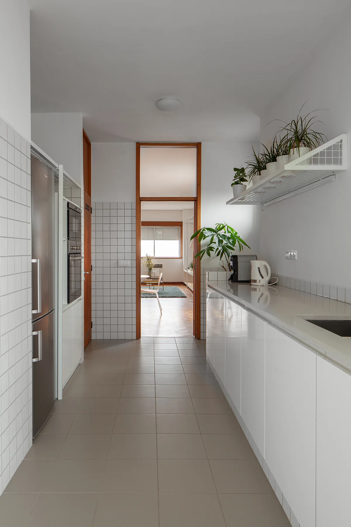 A narrow galley kitchen features glossy white cabinets, a grid of white square wall tiles, gray floor tiles, and a wooden doorframe.