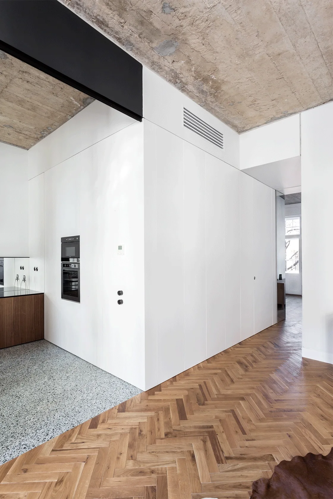 A seamless white kitchen cabinet wall sits between a grey terrazzo floor and warm herringbone wood flooring under an exposed concrete ceiling.