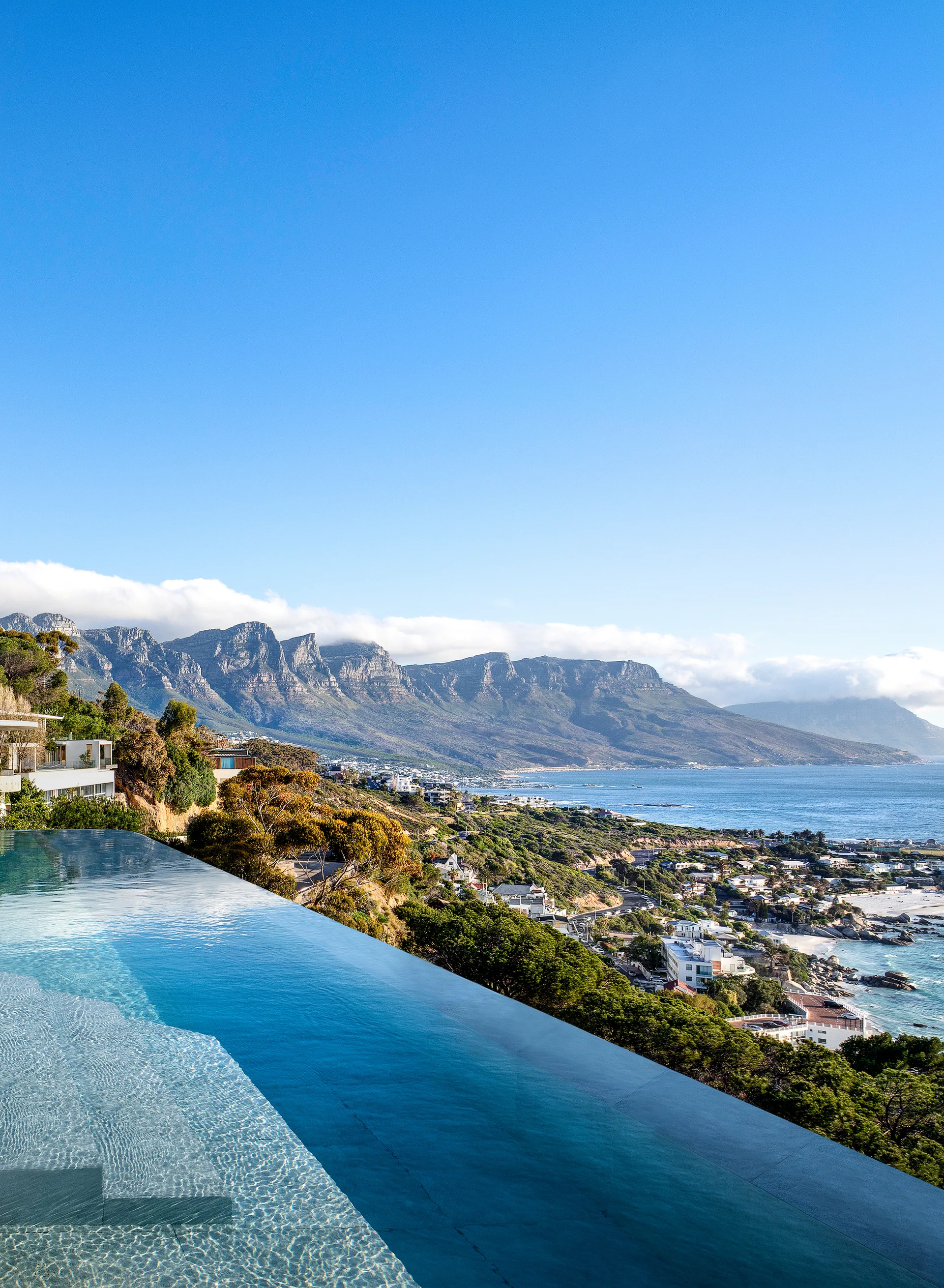 An infinity pool with clear blue water overlooks a coastal landscape of mountains, green hills, and a sandy beach under a clear sky.