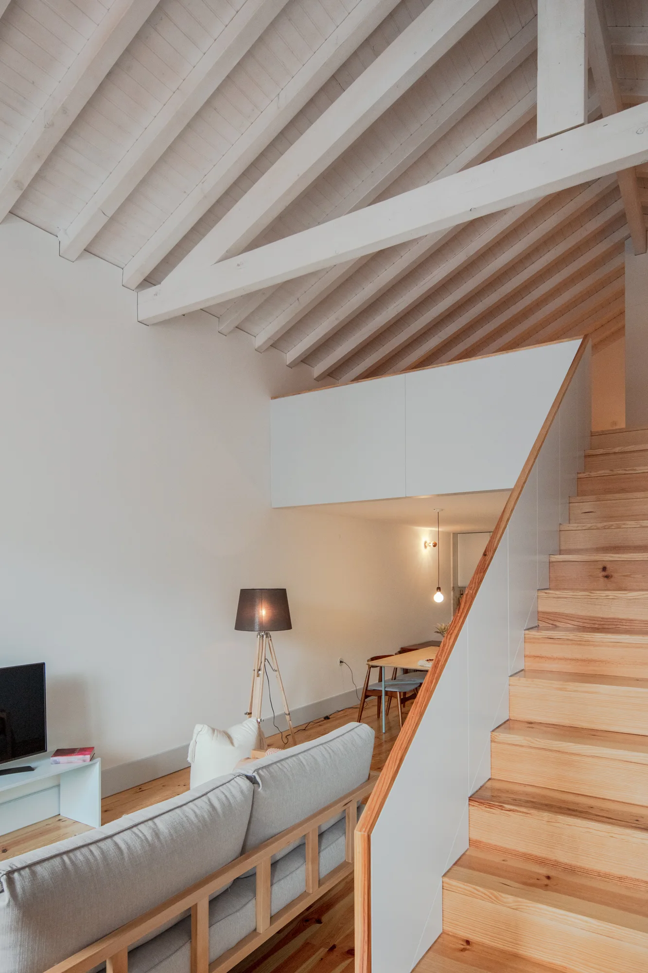A light wood staircase stands next to a grey sofa in a room with white walls and exposed painted ceiling beams.