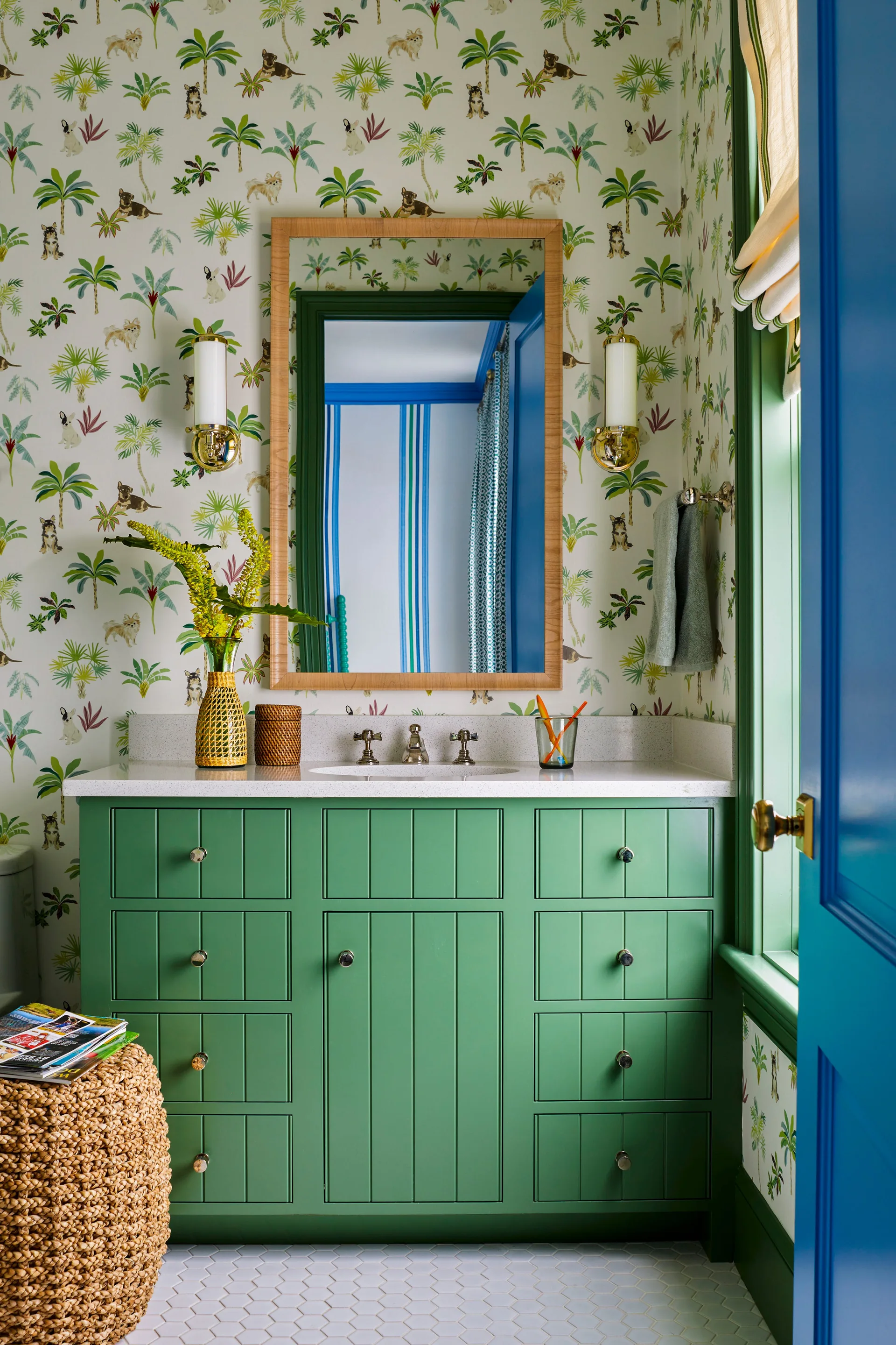 A powder room features a green painted wood vanity, a wood-framed mirror, brass sconces, and whimsical palm-and-dog patterned wallpaper.