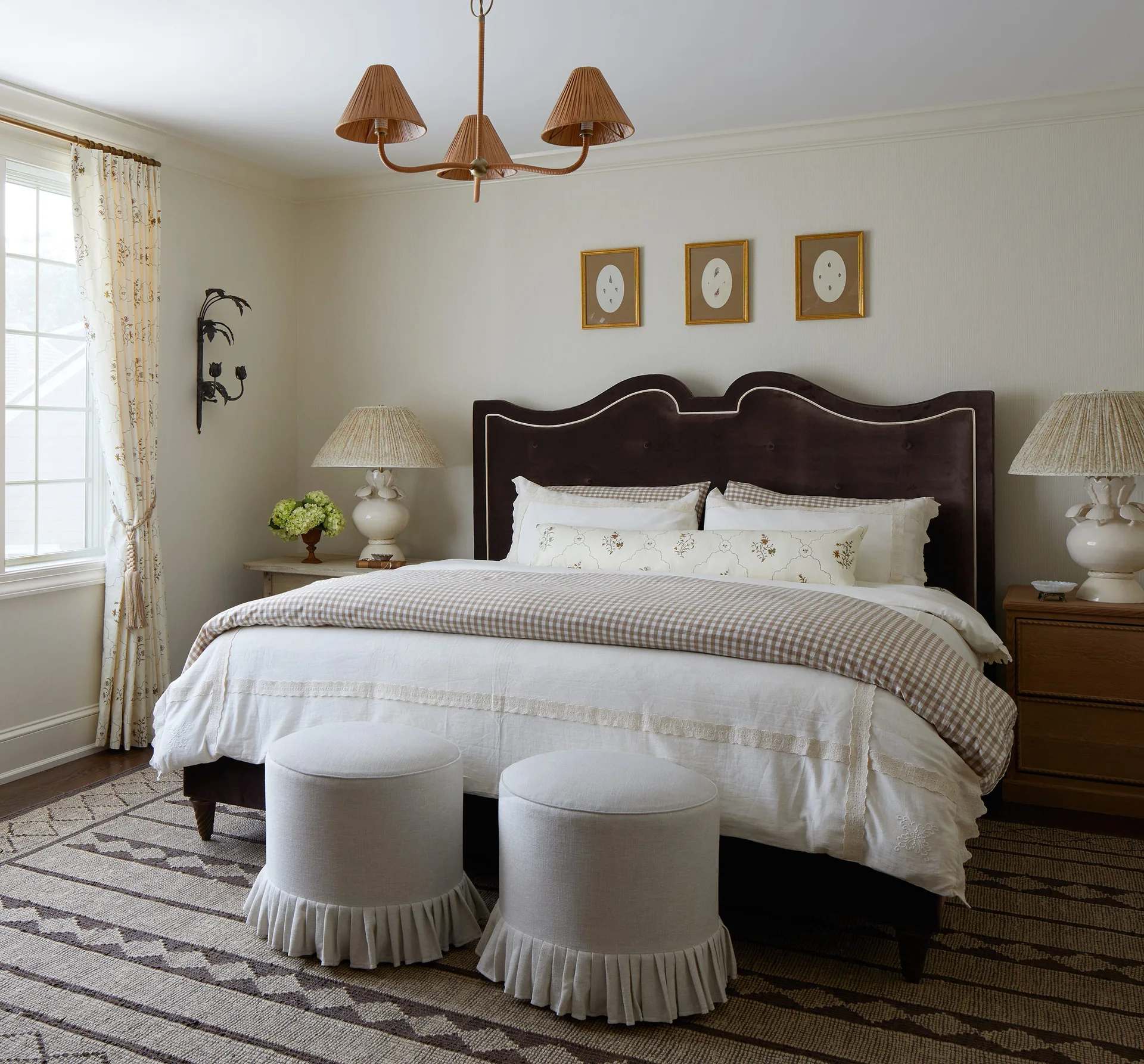 A traditional bedroom features a dark scalloped headboard, gingham bedding, and two matching round skirted ottomans at the foot of the bed.