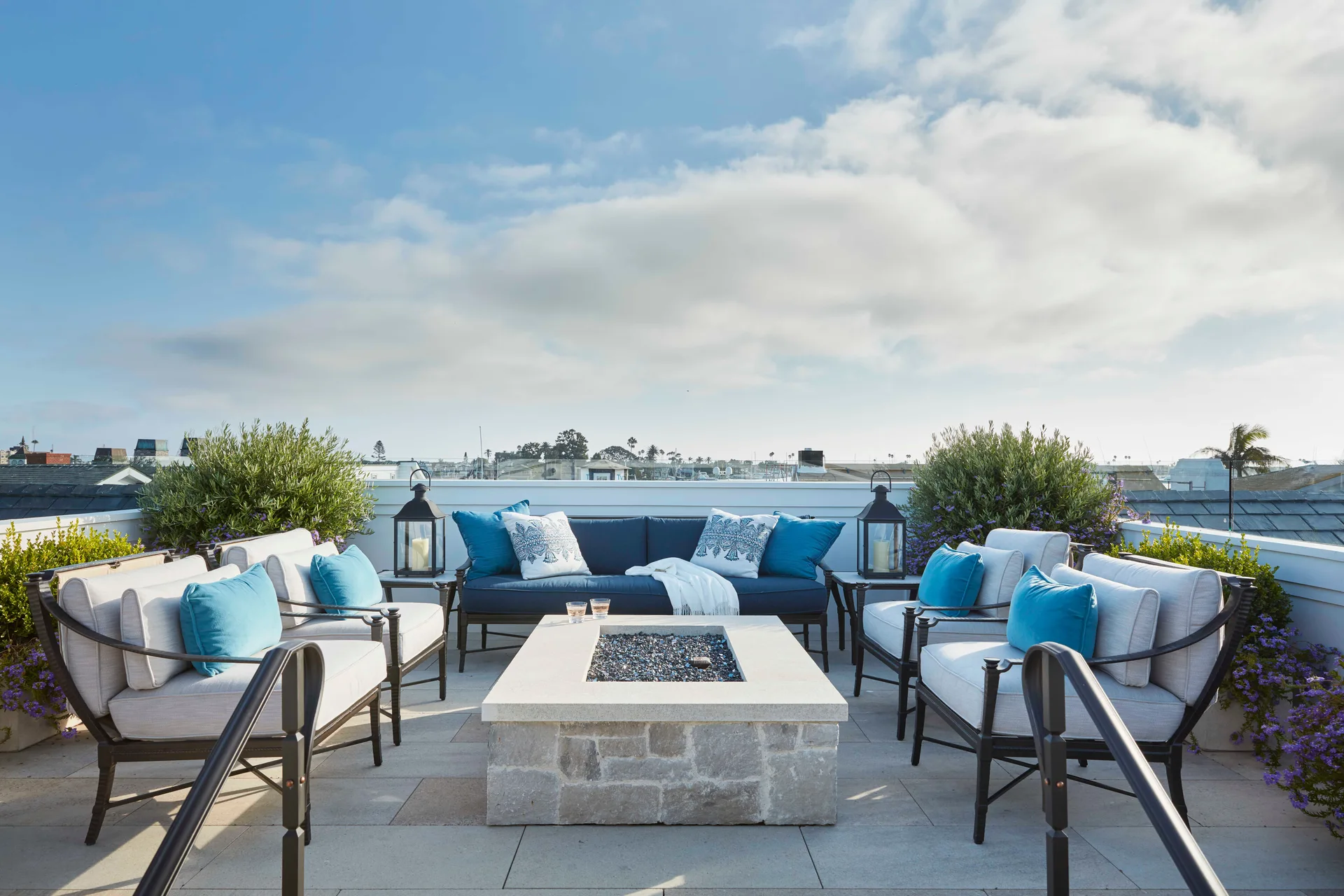 A rooftop terrace seating area featuring a central rectangular stone fire pit surrounded by a blue sofa and white cushioned armchairs.