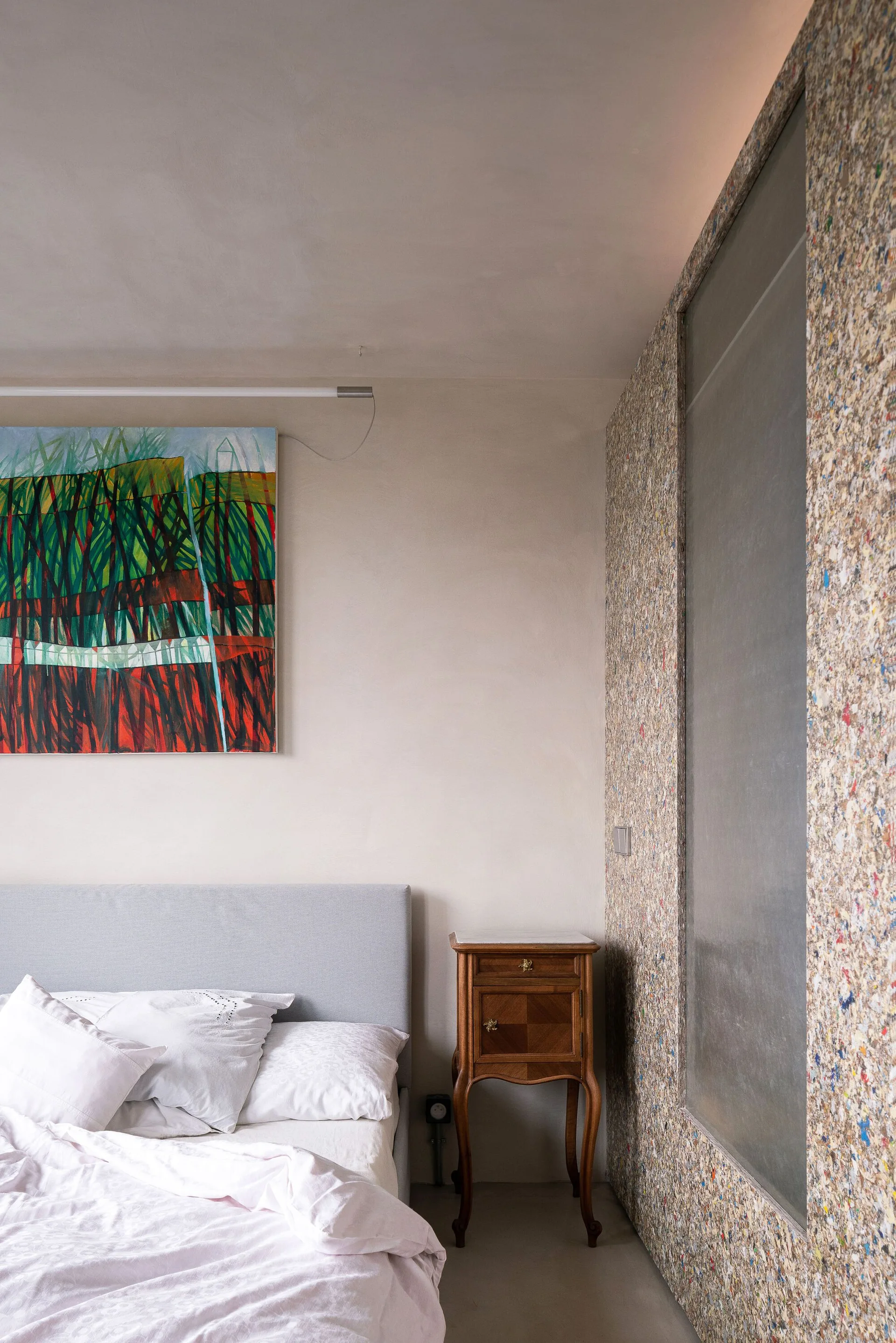 A modern bedroom featuring a gray upholstered bed, a vintage wooden nightstand, a colorful painting, and a speckled composite accent wall partition.