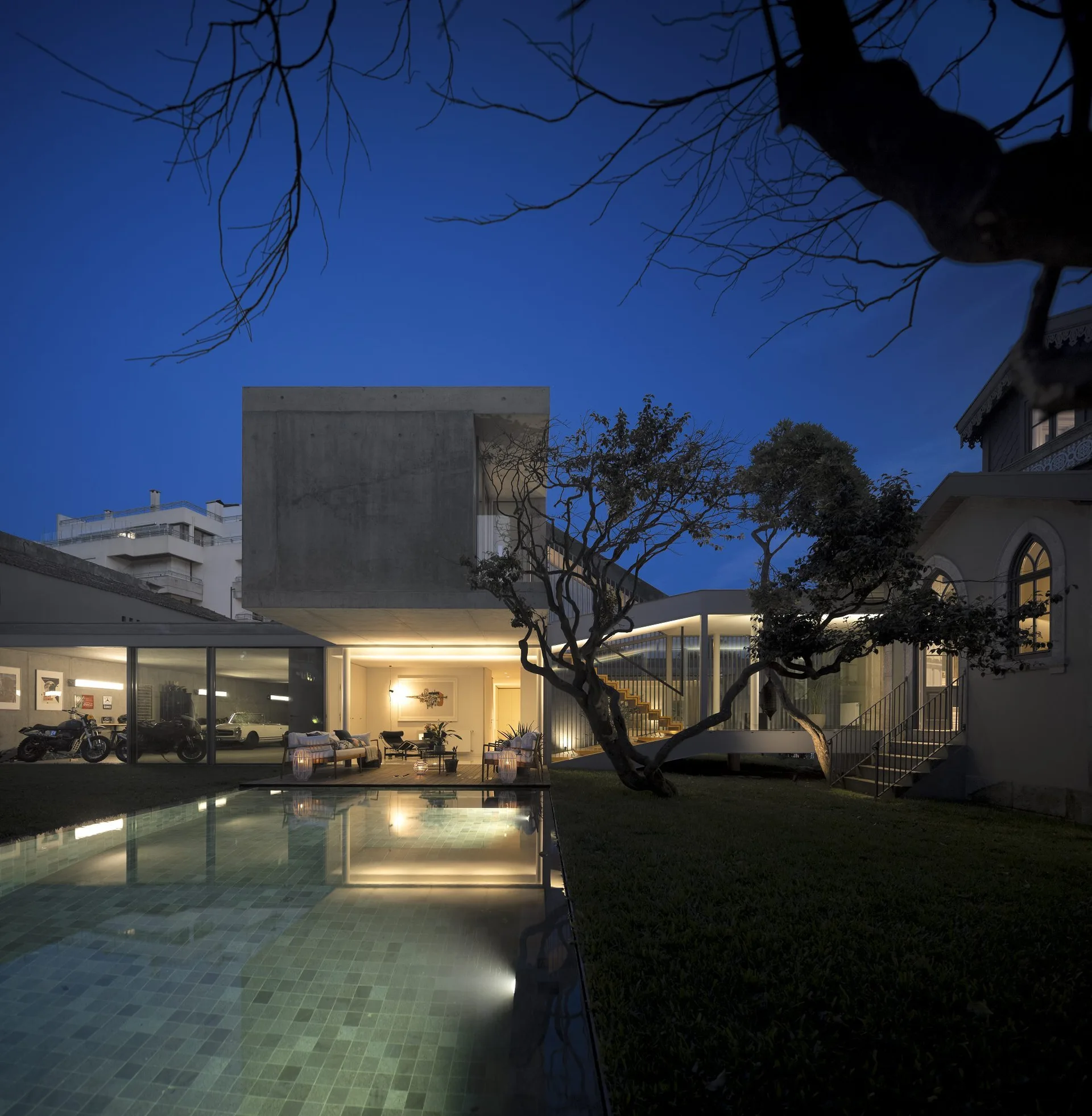 A contemporary concrete house with floor-to-ceiling glass walls is illuminated at dusk next to a swimming pool and a wooden deck.