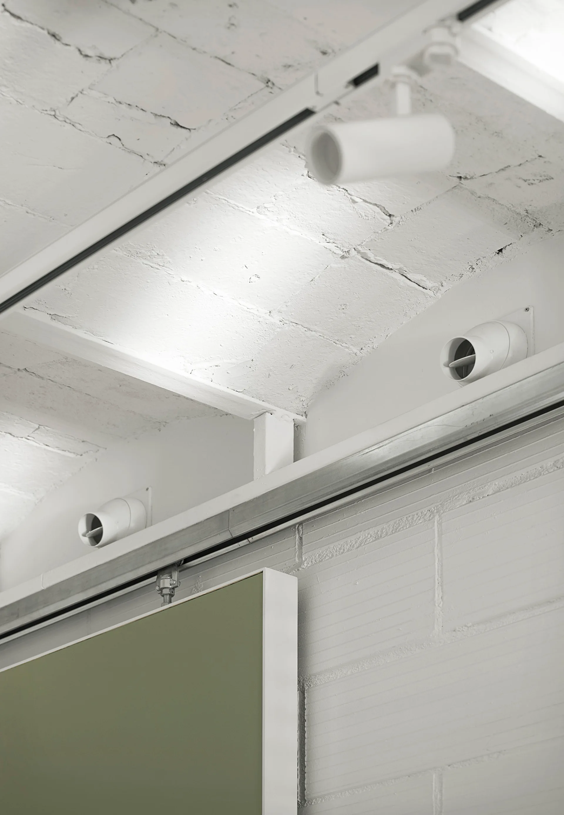 An exposed white brick ceiling features modern track lighting and circular vents above a matte olive green sliding wall panel.