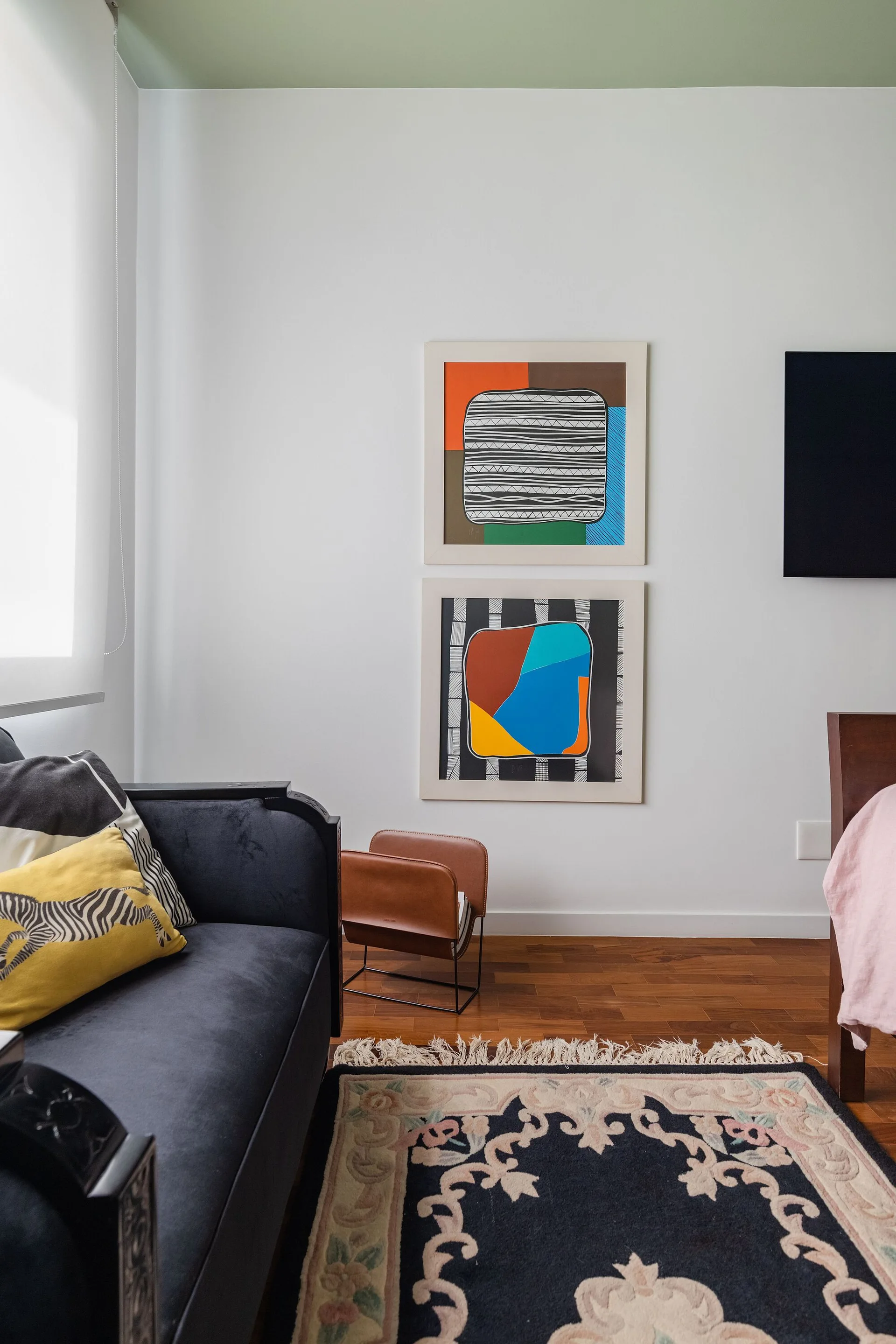 An eclectic bedroom corner features a dark blue sofa, patterned rug, leather magazine holder, and stacked art below a green painted ceiling.