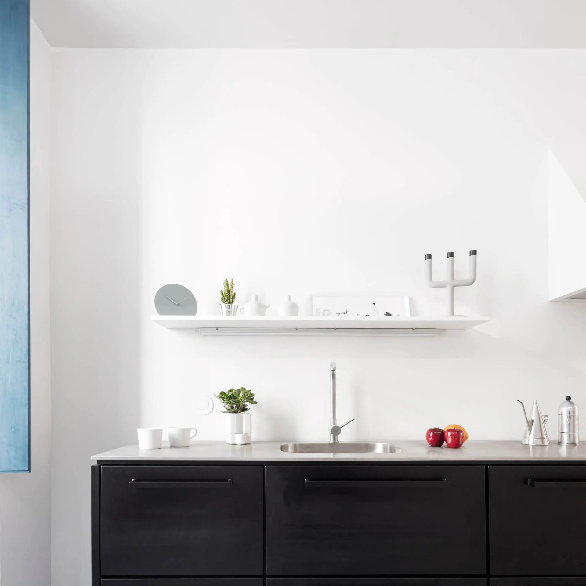 Matte black lower cabinets and a stainless steel sink are set against a plain white wall featuring a single floating shelf with minimalist decor.
