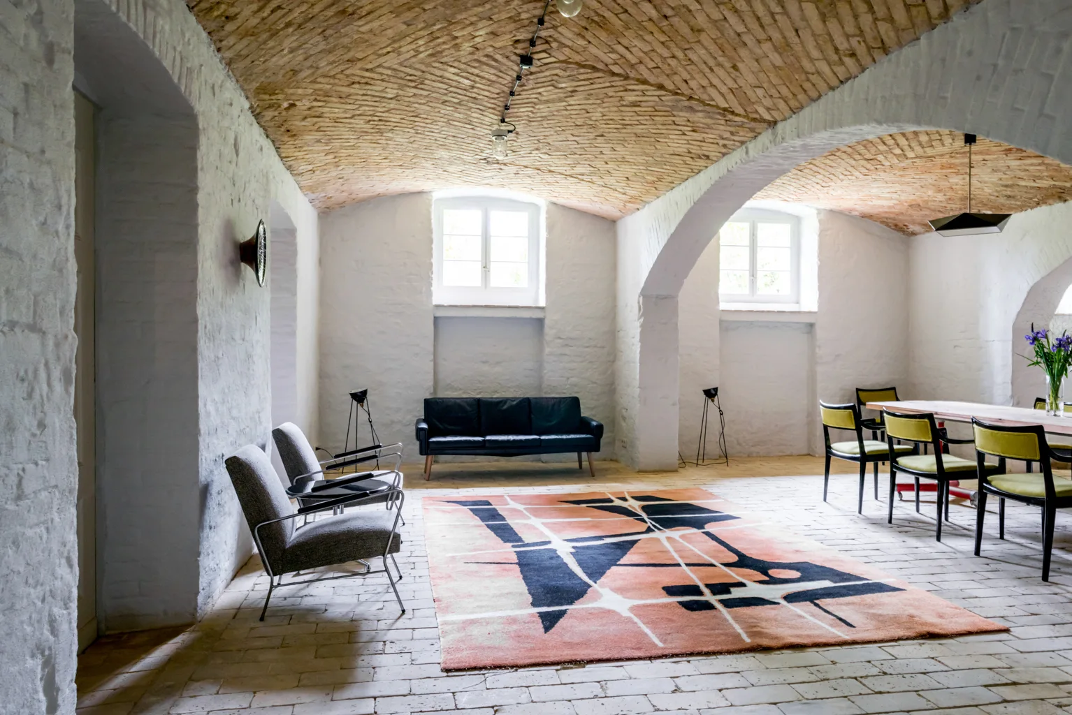 An open-concept room features a curved brick vaulted ceiling, white painted brick walls, a bold geometric rug, and mid-century modern furniture.
