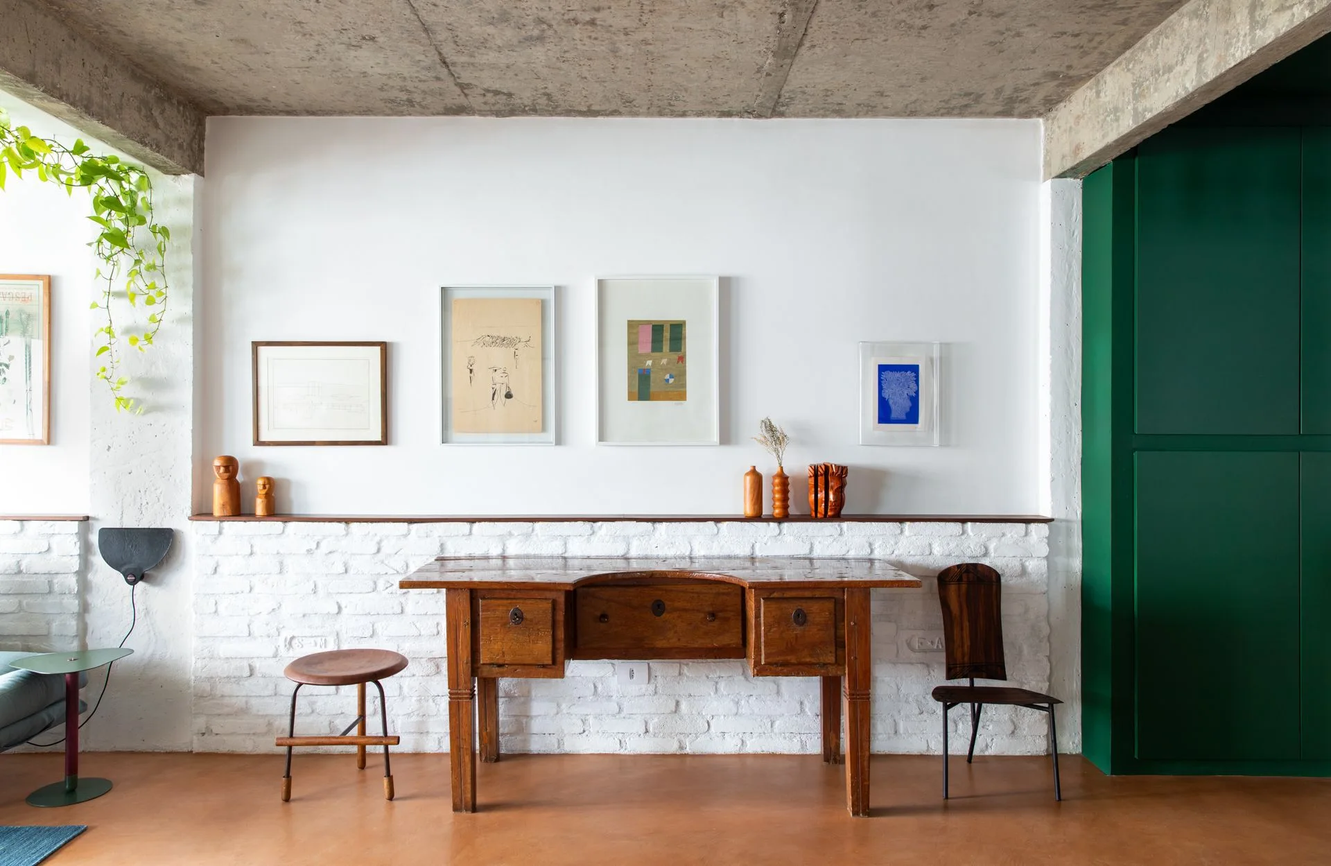An antique wooden desk sits against a white painted brick wall under a concrete ceiling, flanked by a wooden stool and chair.
