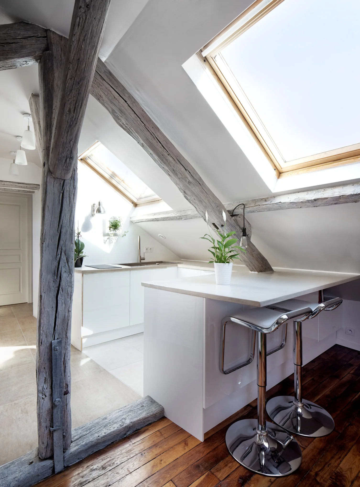 A bright attic kitchen features sloped ceilings with skylights, weathered wooden beams, and a modern white island with two chrome bar stools.