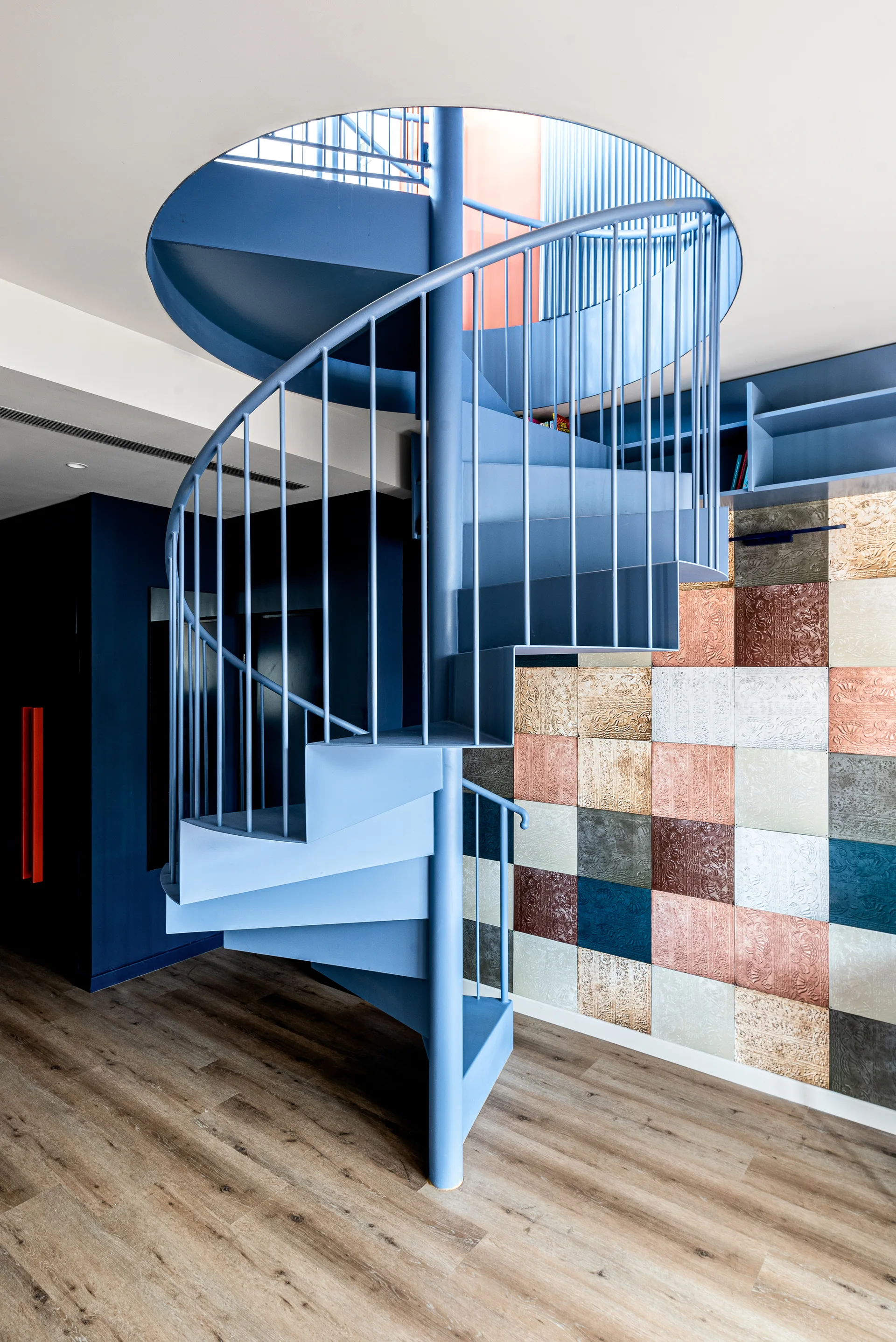 A matte blue metal spiral staircase winds up through a circular ceiling opening next to a wall clad in colorful embossed metallic tiles.