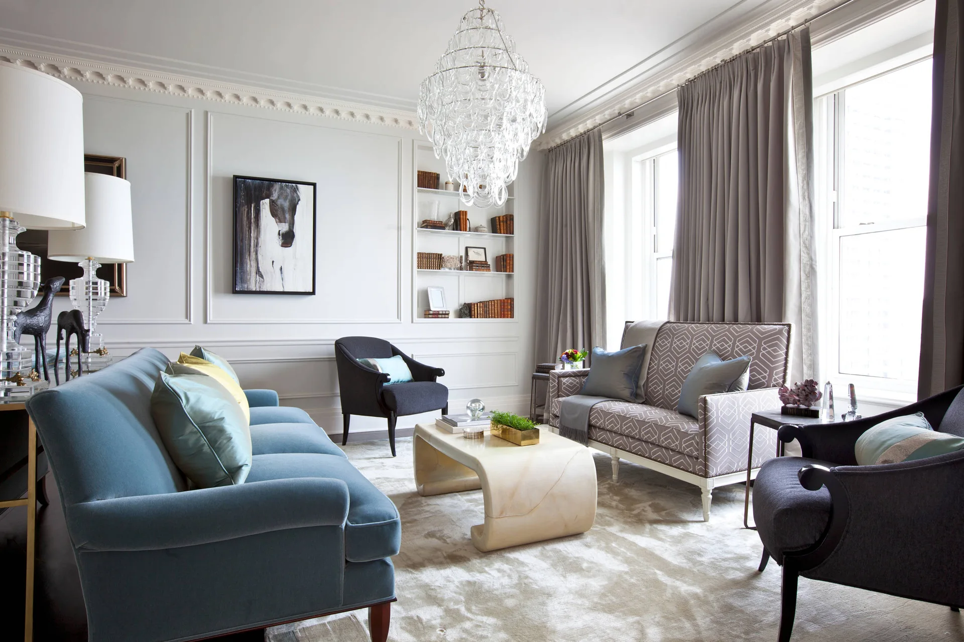A transitional living room features a blue velvet sofa, a curved stone coffee table, and a crystal chandelier beneath decorative crown molding.