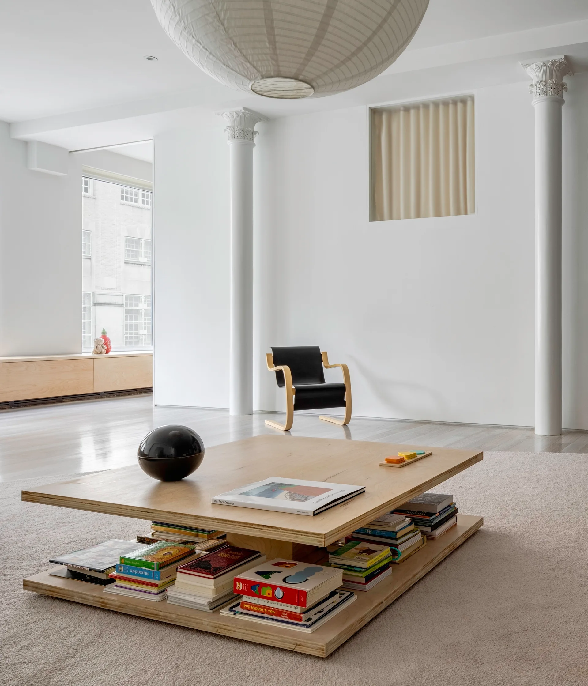 A minimalist living room features a low two-tier plywood coffee table stacked with books, a black lounge chair, and white fluted classical columns.