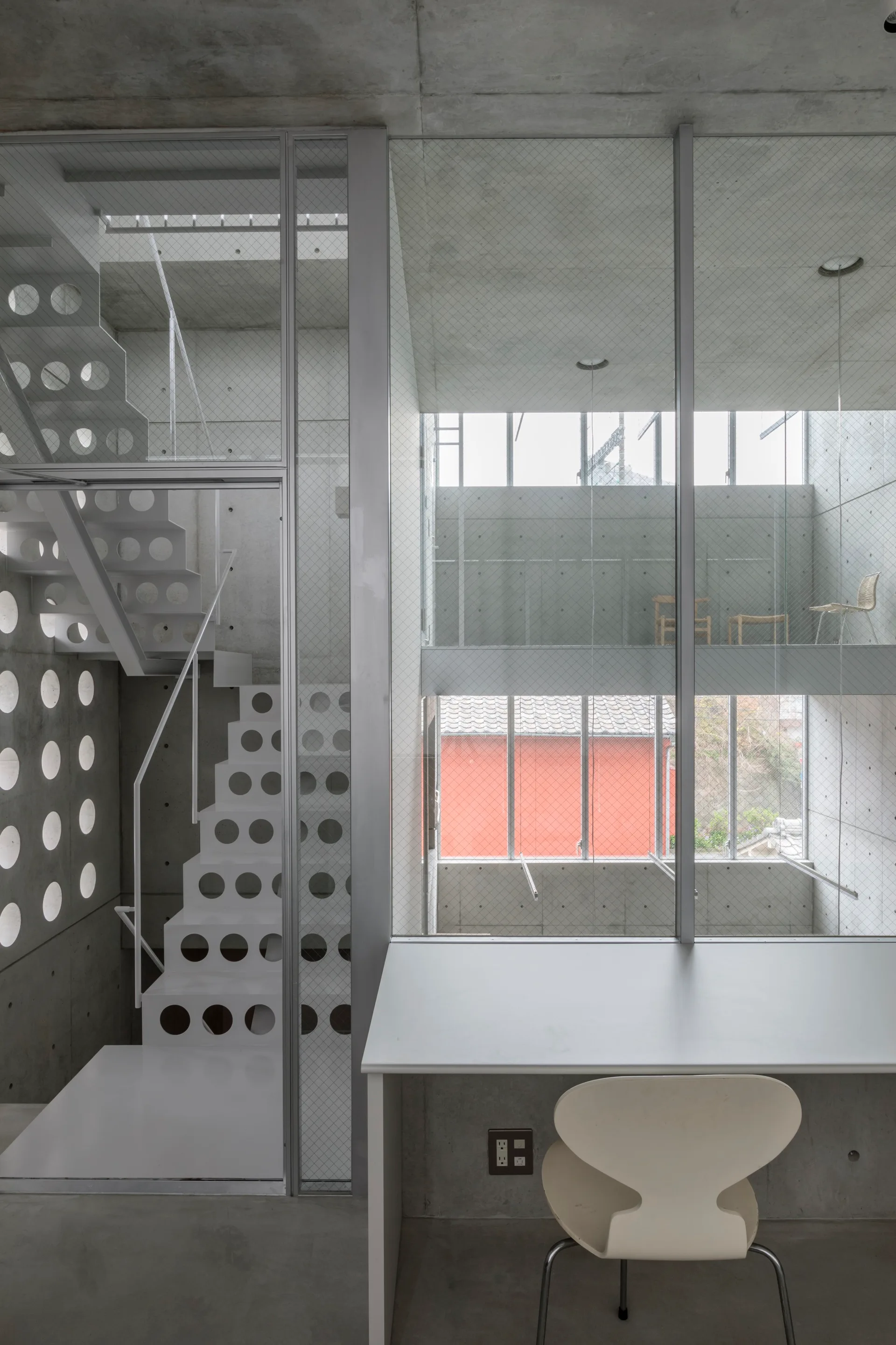 A minimalist home office features a white curved chair at a desk, adjacent to a white perforated metal staircase and exposed concrete walls.