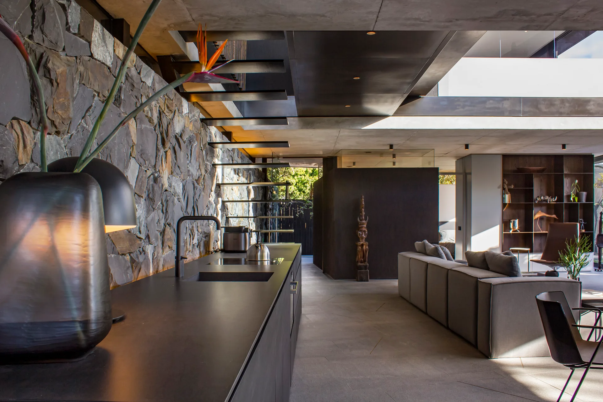 A modern open-plan kitchen and living room featuring a textured stone feature wall, floating stairs, a black island, and a grey sectional sofa.