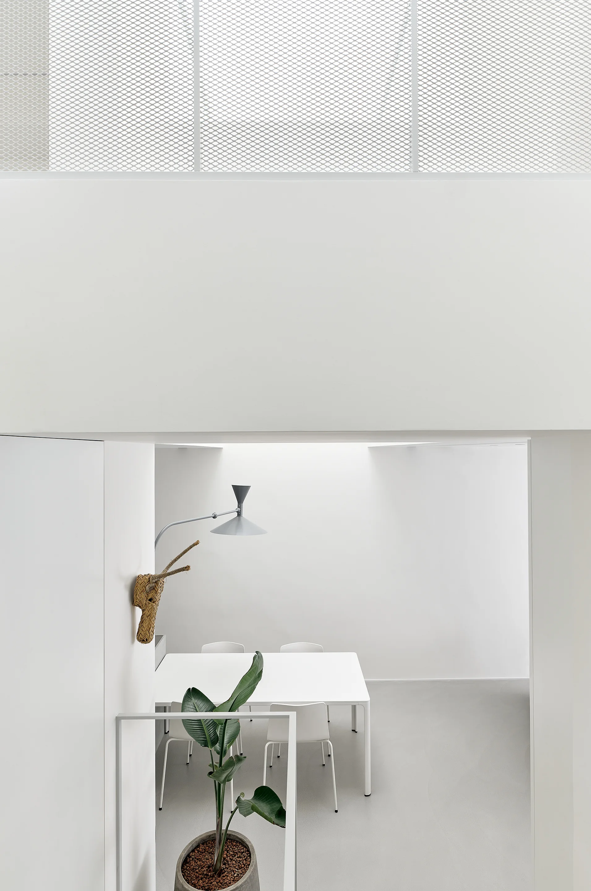 A minimalist white dining room with a simple table, chairs, a grey swing-arm sconce, and a green potted plant viewed from an upper level.