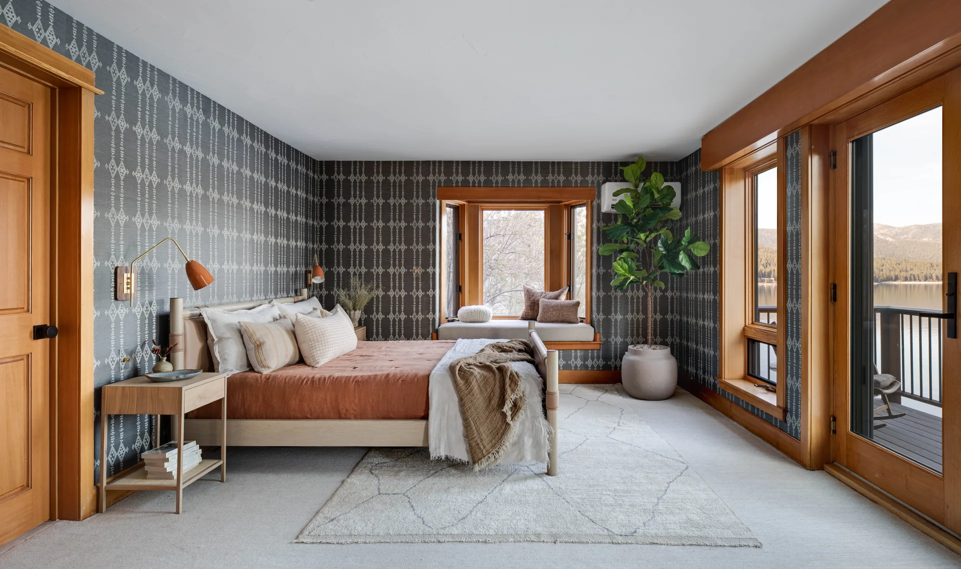 A modern bedroom features charcoal geometric wallpaper, a light wood bed with terracotta bedding, and warm wood-framed windows looking out to a lake.