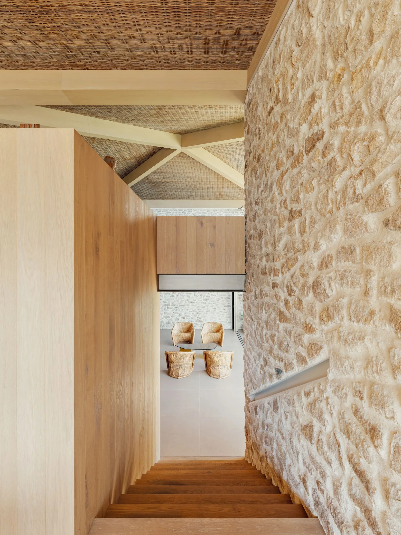 A wooden staircase descends next to a rough stone wall, looking down toward a seating area with four wicker armchairs and a woven ceiling.
