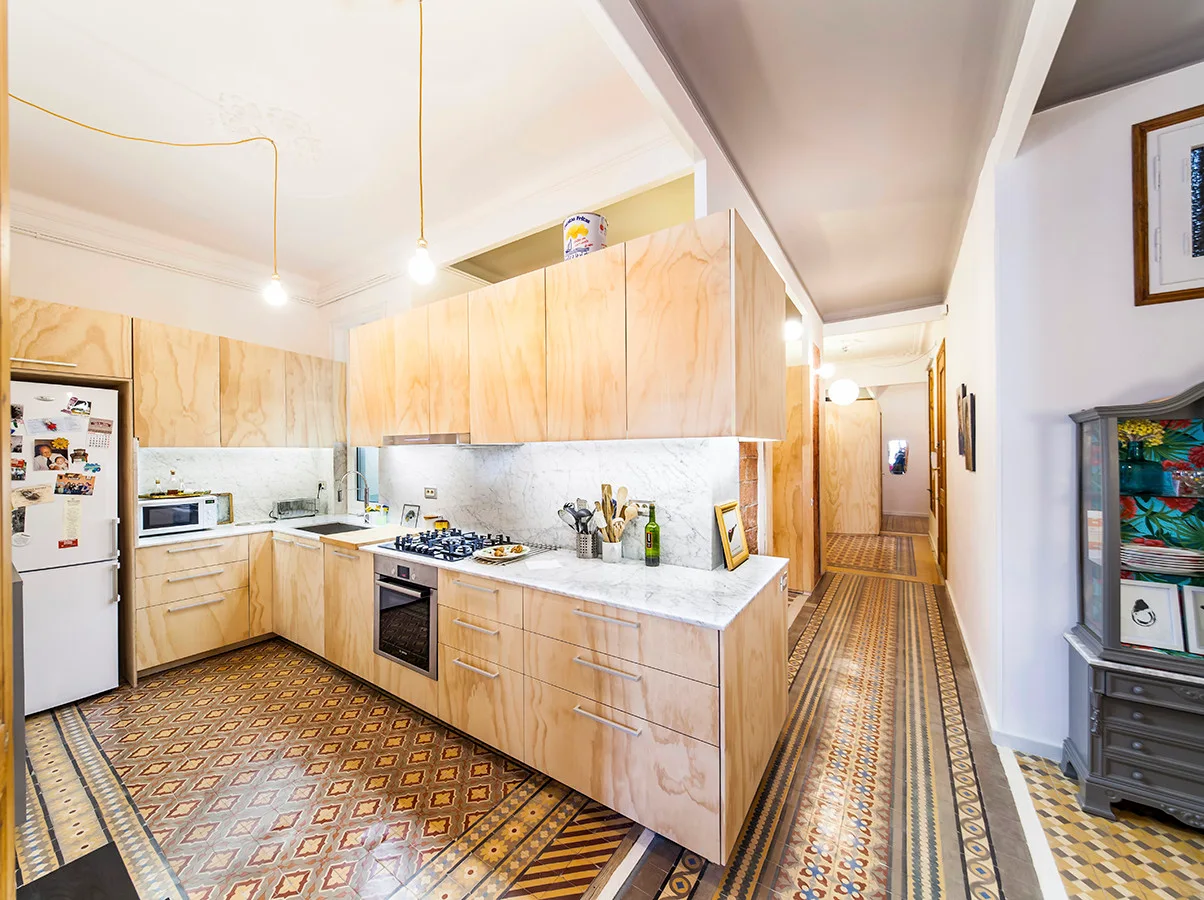 A residential kitchen features light plywood cabinetry, white marble countertops, built-in appliances, and a highly patterned, colorful tile floor.