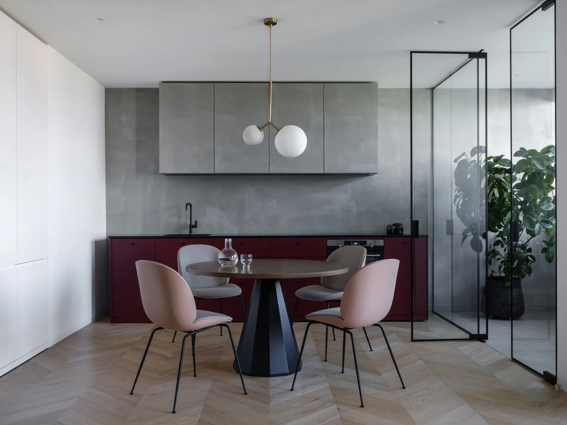 A modern kitchen and dining space features burgundy lower cabinetry, a round wooden table with pink and gray chairs, and glass partition doors.
