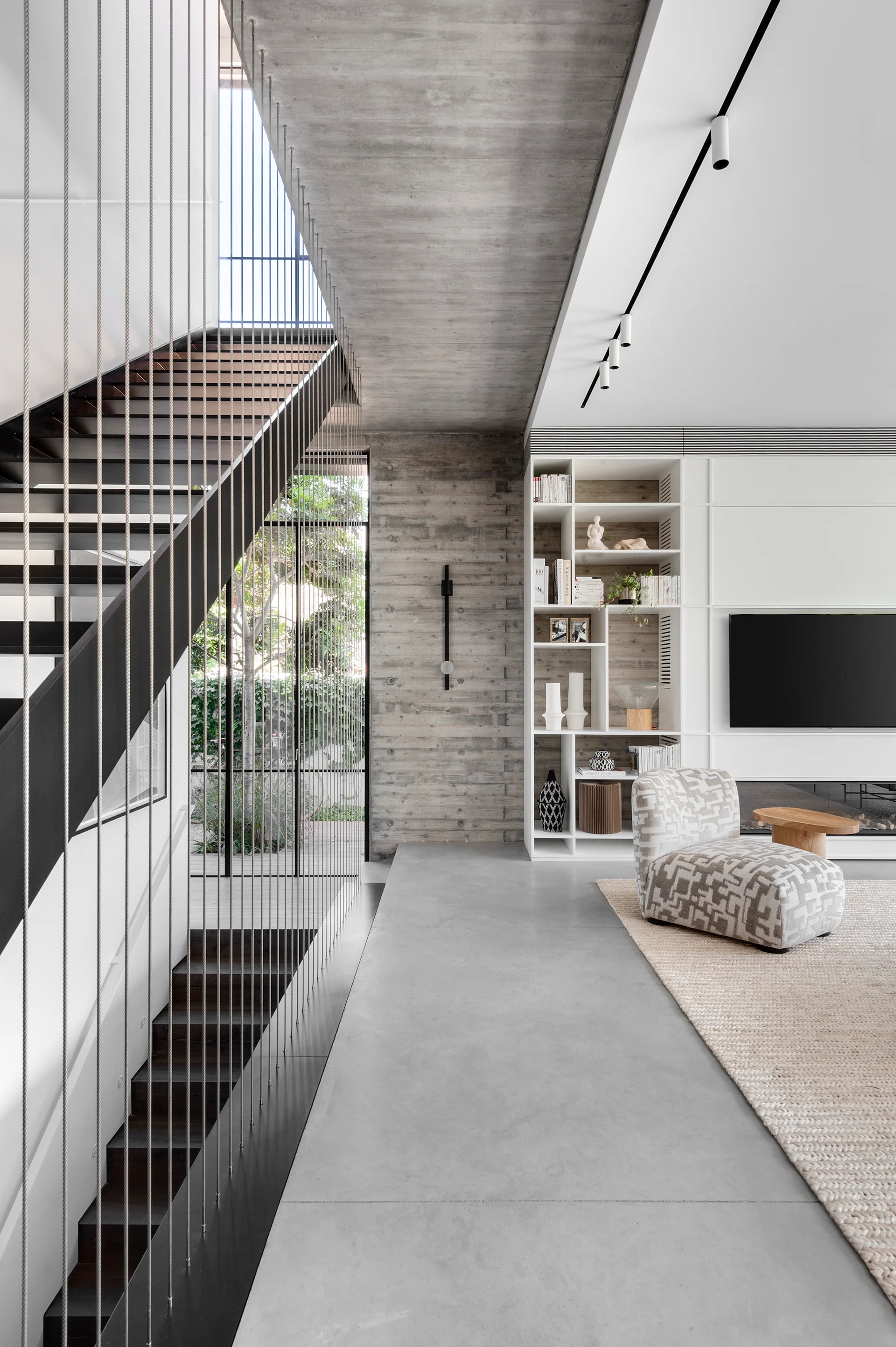 A modern living room features a staircase with vertical cable railings next to a board-marked concrete wall and white built-in shelving.