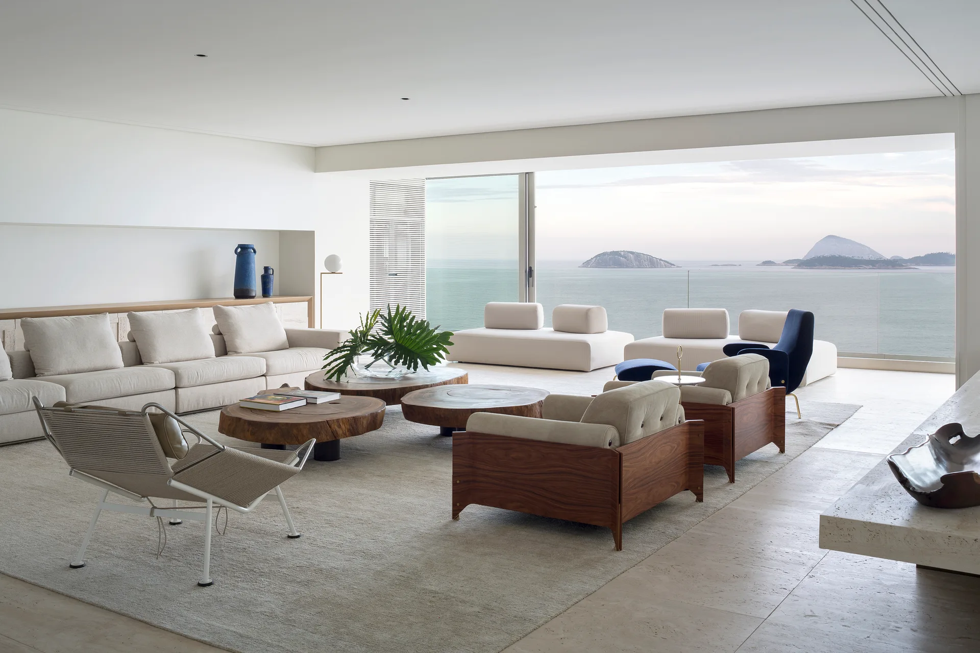 A minimalist coastal living room features low-profile beige sofas, organic wood-slab coffee tables, and a large sliding glass door opening to an ocean view.